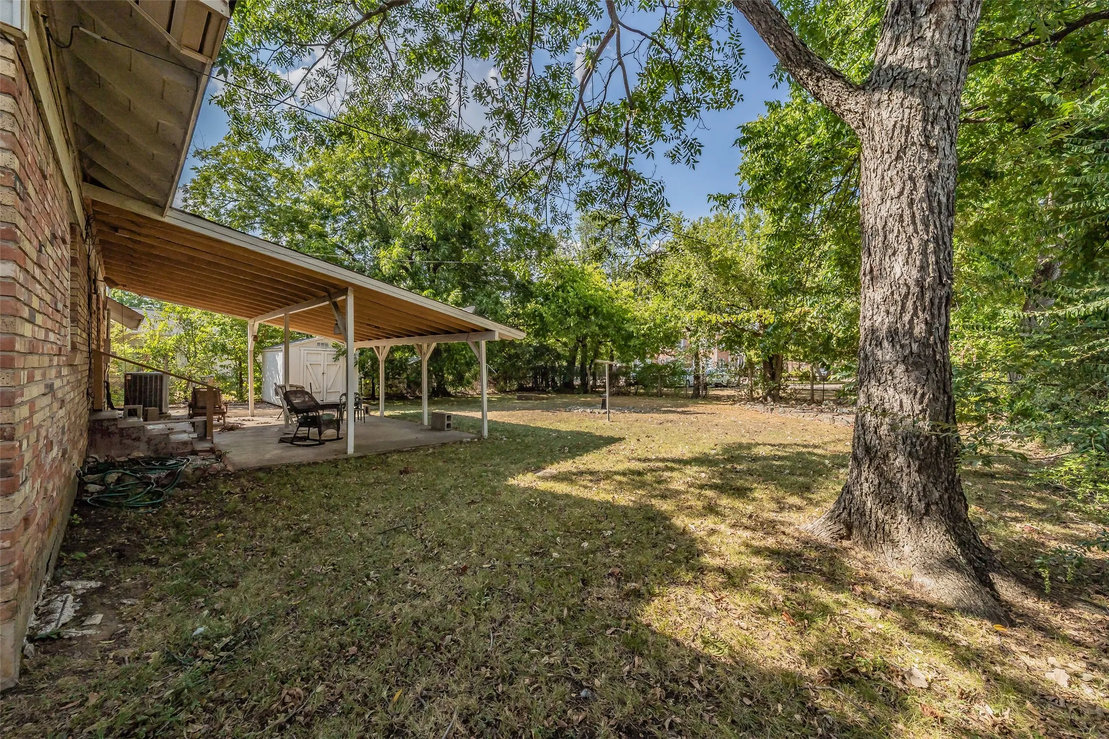 Expansive backyard featuring a covered patio with concrete flooring, mature trees, and a brick exterior