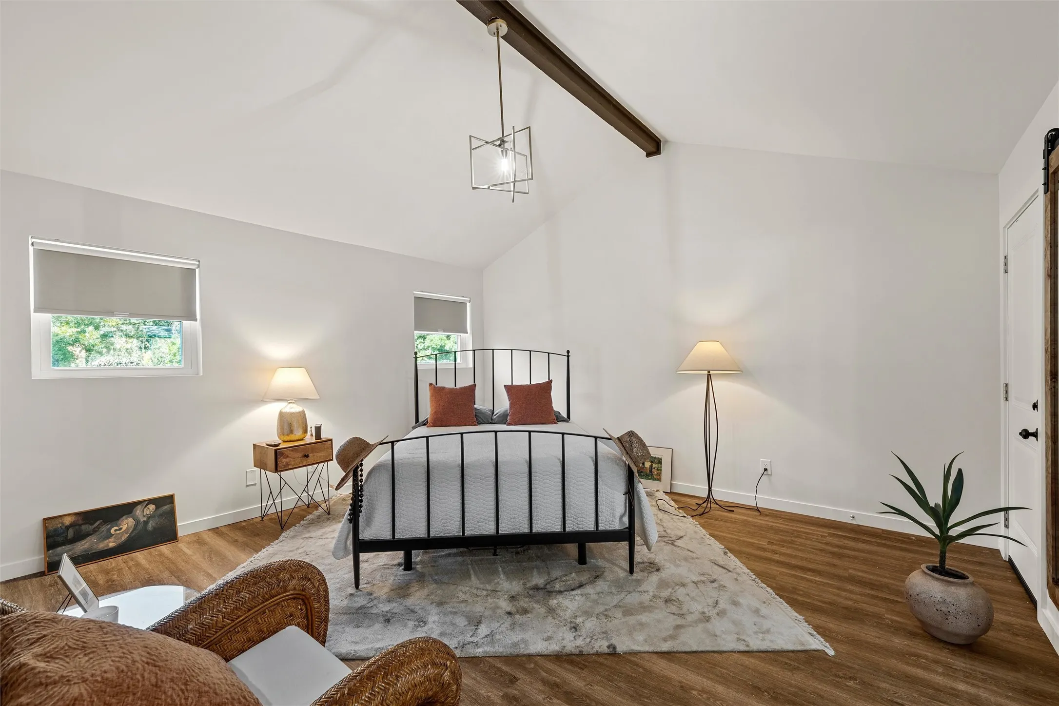 Vaulted ceilings with an exposed beam, wood-finish flooring, two windows with roller shades, and a white barn door