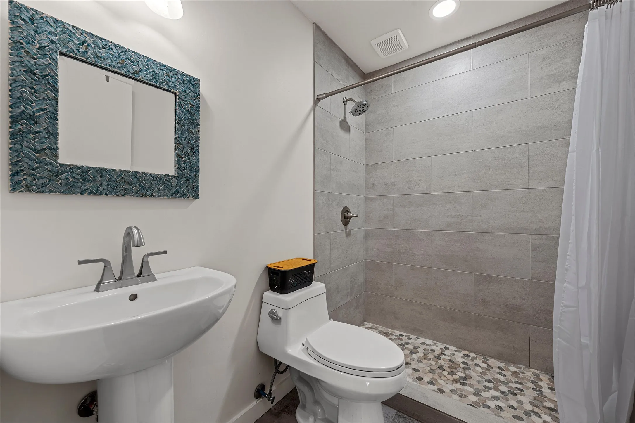 Pedestal sink with chrome faucet, textured mirror, tiled shower walls, pebble shower floor, and recessed lighting