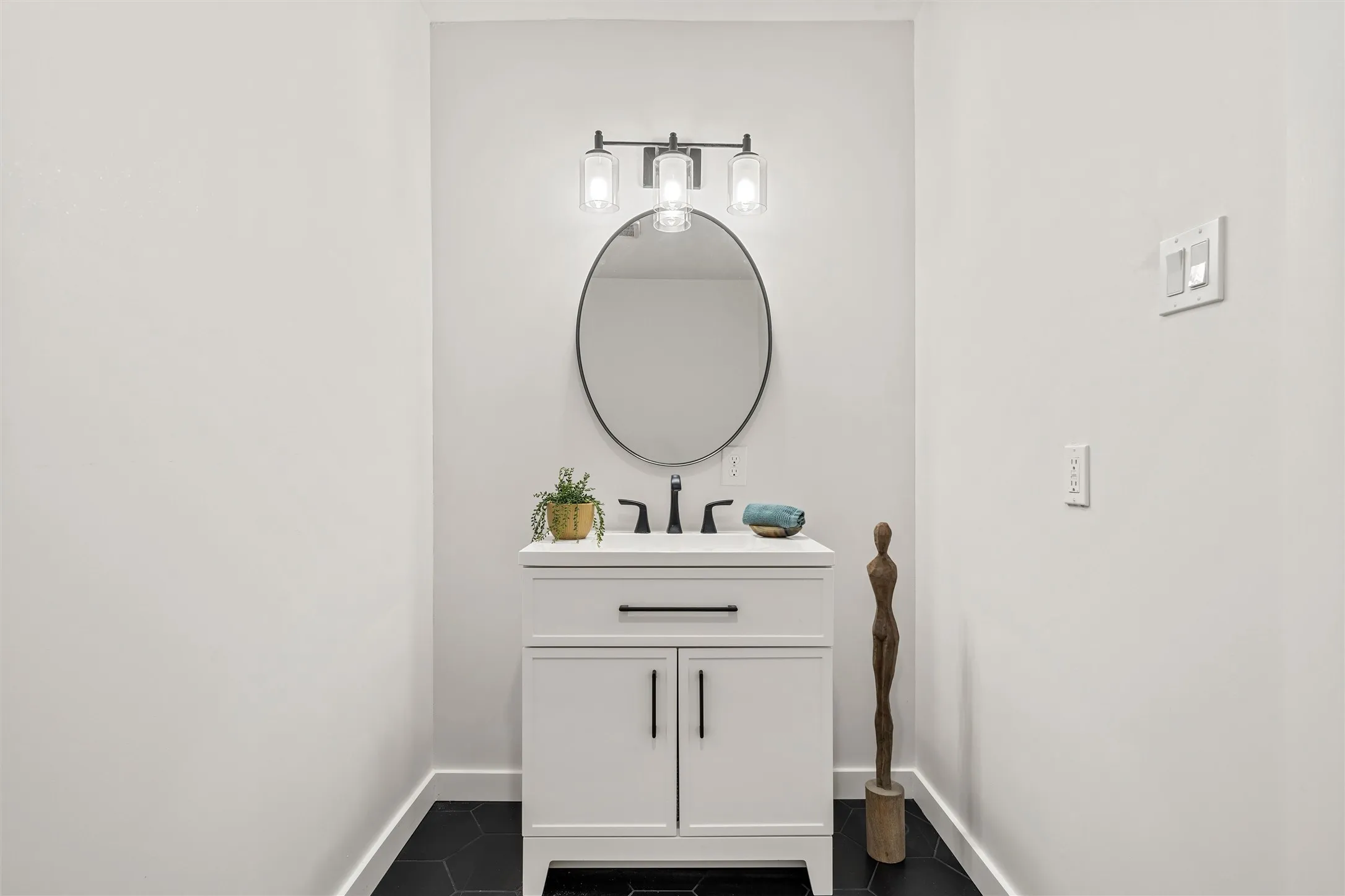 White vanity with matte black hardware, an oval mirror, a three-light vanity fixture, and hexagonal floor tiling
