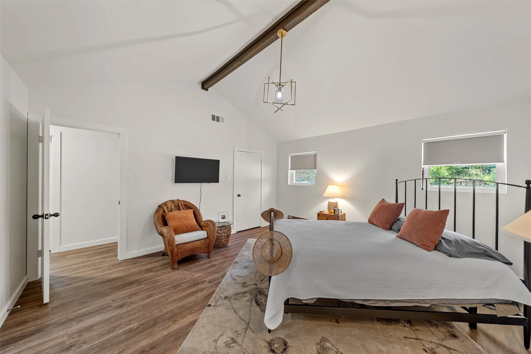 Vaulted ceiling with exposed wood beam, wood-finish flooring, two windows with roller shades, and a geometric pendant light fixture