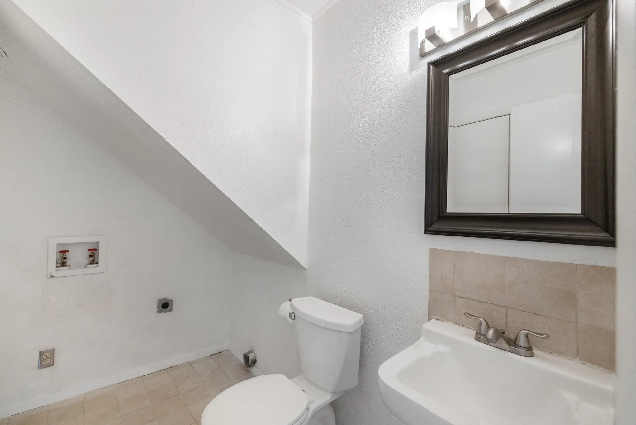 White ceramic toilet, pedestal sink with dual-handle faucet, framed mirror, and tiled​​‌​​​​‌​​‌‌​‌‌​​​‌‌​‌​‌​‌​​​‌​​ backsplash