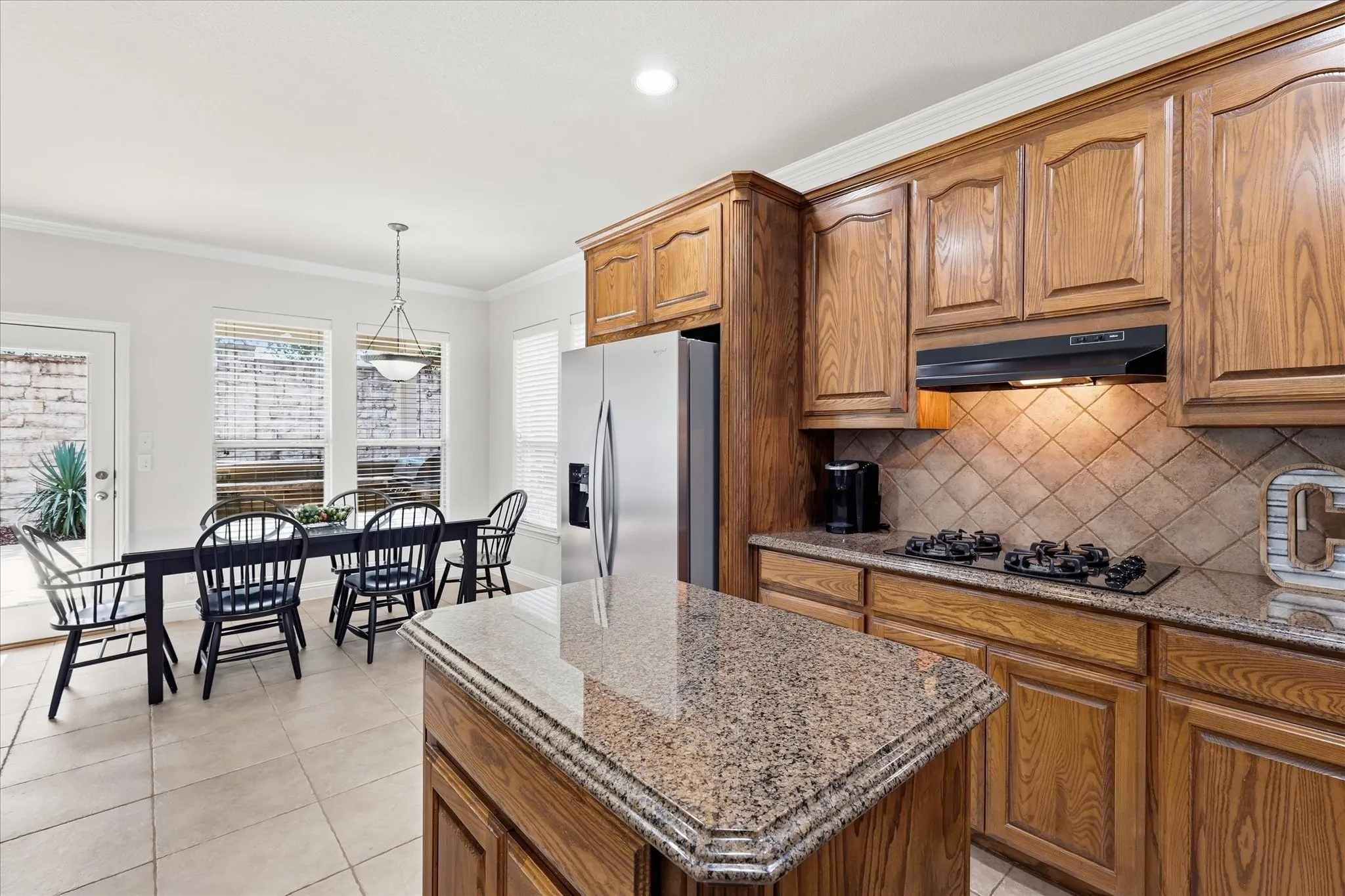 Kitchen featuring wood cabinetry, granite countertops, tile flooring, stainless steel refrigerator, and built-in cooktop with tile backsplash
