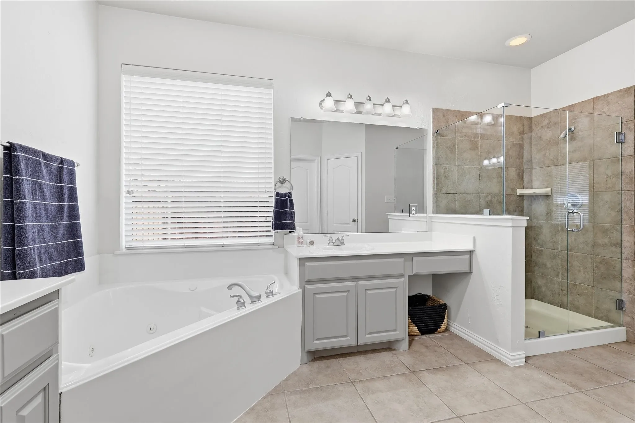 Corner soaking tub, separate glass-enclosed shower with tile surround, double vanity with gray cabinetry, white countertops, and tiled flooring