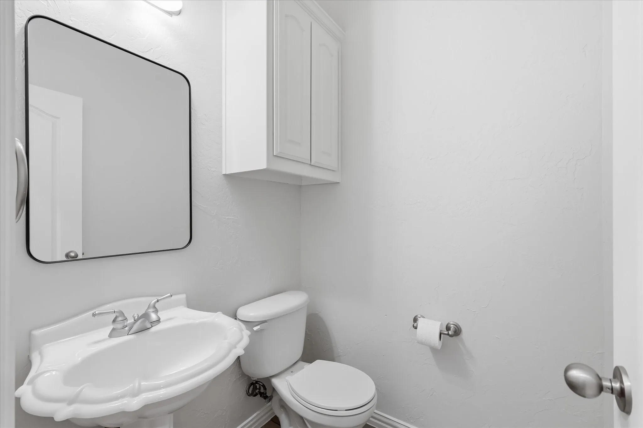 Pedestal sink with chrome fixtures, wall-mounted mirror, white toilet, and white upper cabinetry