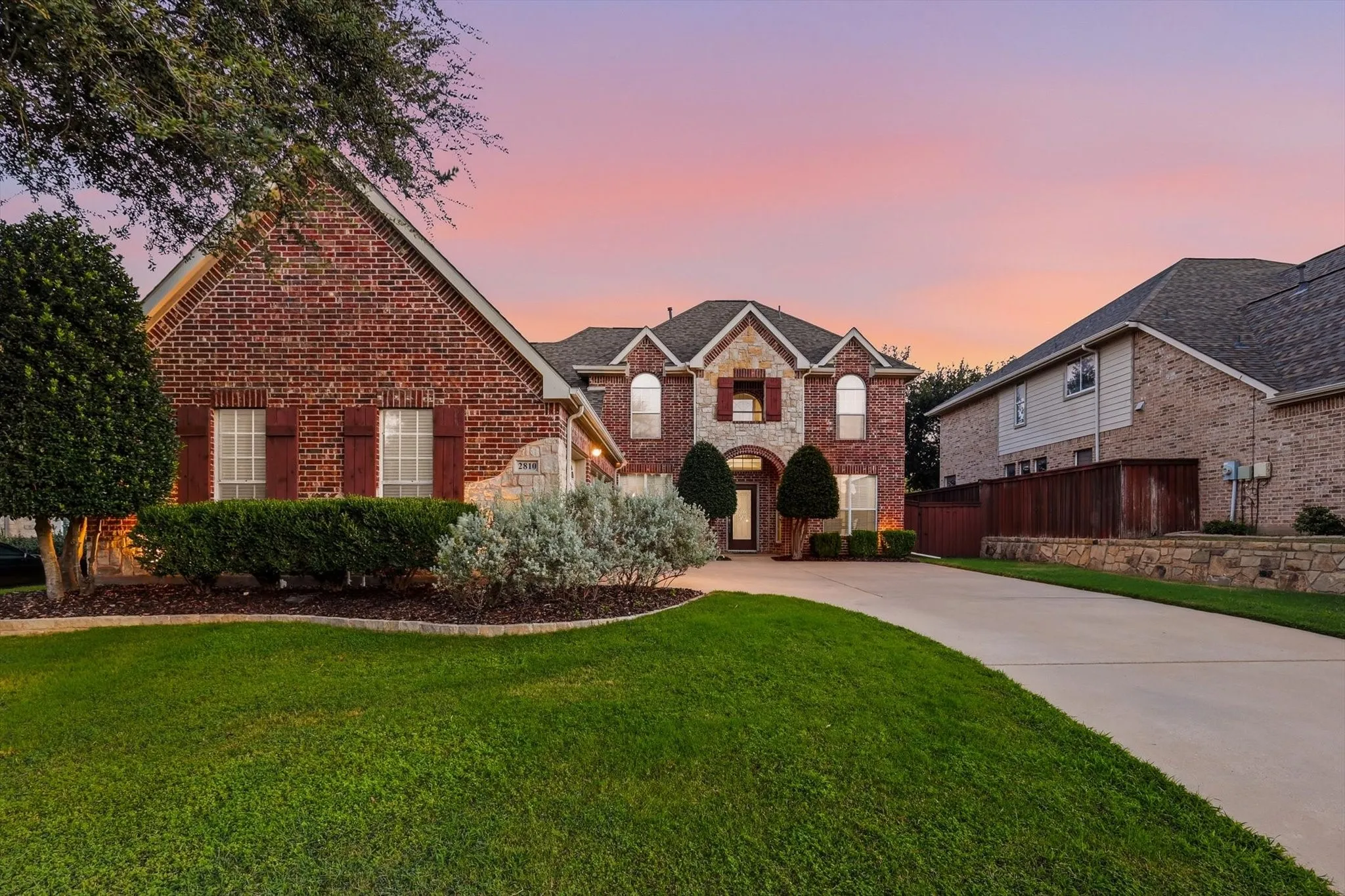 Red brick facade with stone accents, mature landscaping, expansive concrete driveway, and gabled roofline