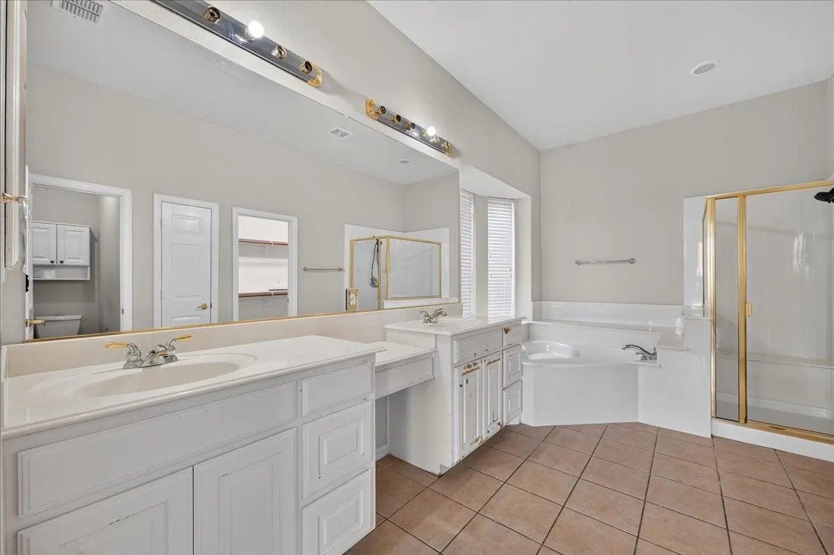 Dual vanity with integrated sinks, large wall mirror, built-in soaking tub, separate shower enclosure with gold-tone framing, and tiled flooring