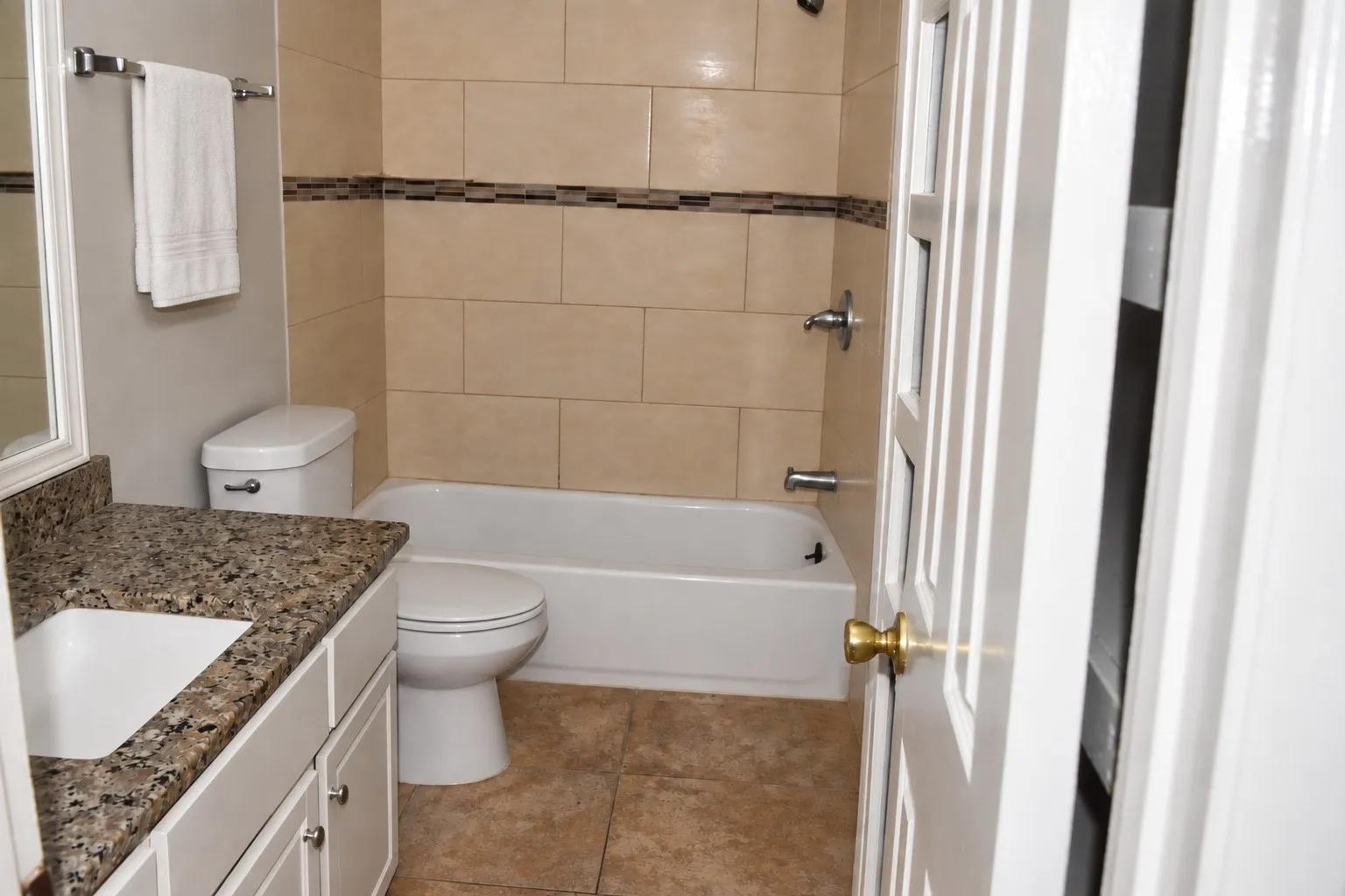 Floor-mounted toilet, built-in bathtub, light-toned wall tiling with decorative accent band, integrated sink, and vanity with stone-finish countertop