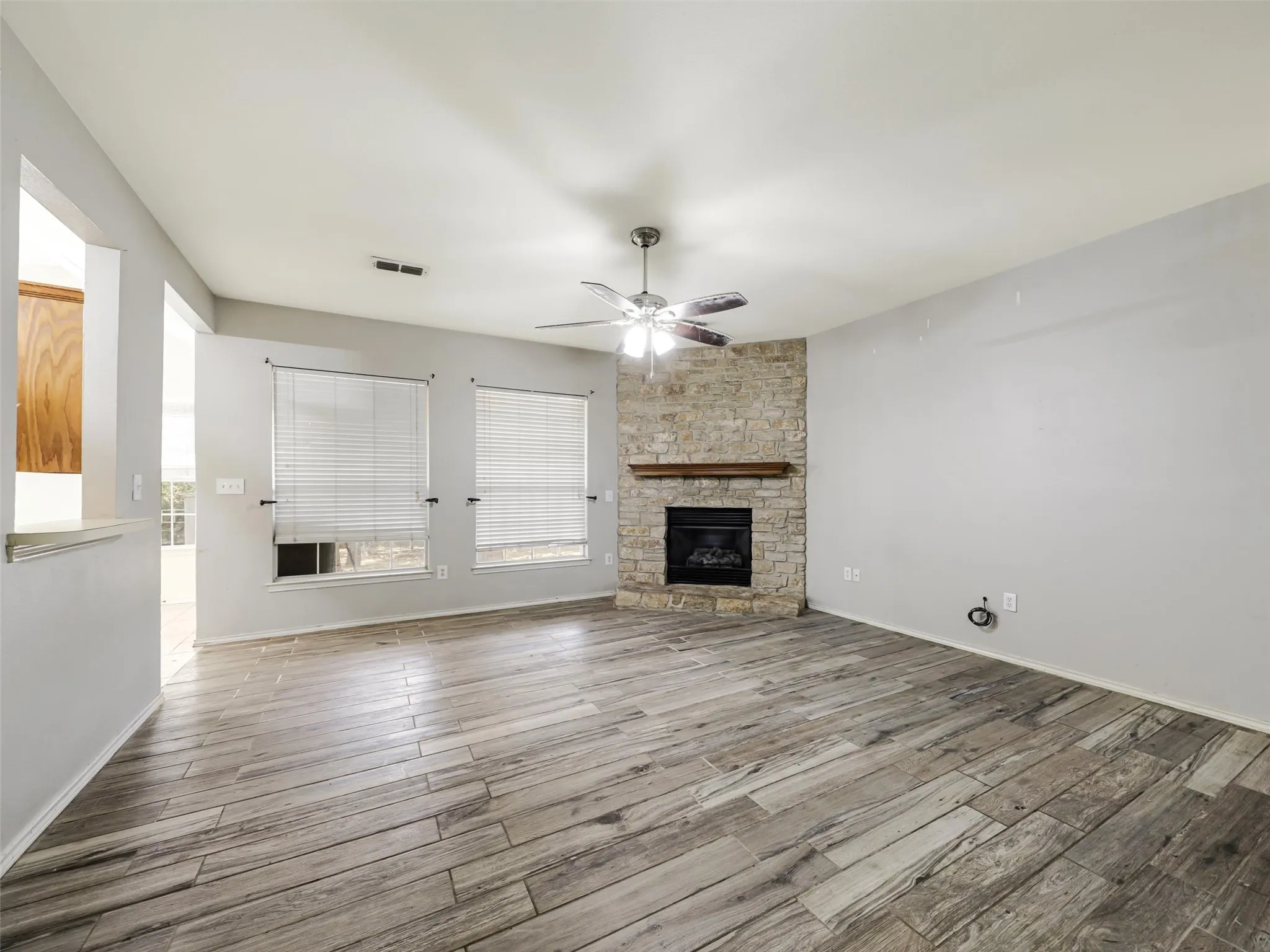 Wood-finish flooring throughout, corner stone-faced fireplace with wood mantel, ceiling fan, and two large windows with blinds