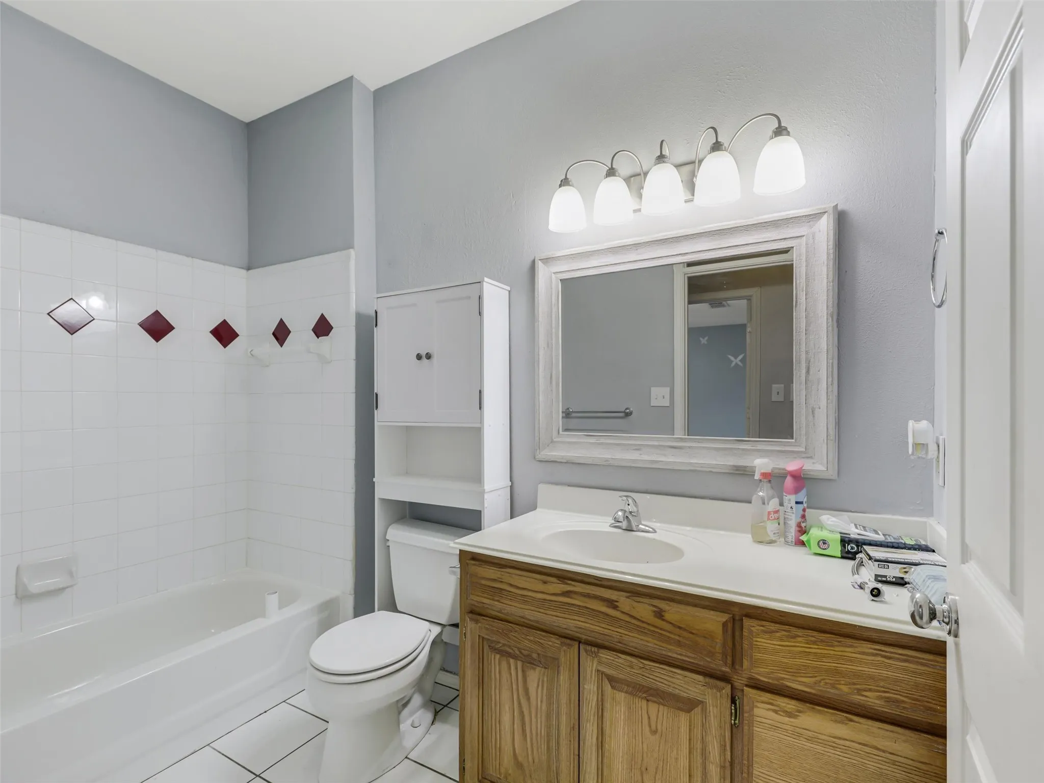 Bathroom featuring a vanity with a white countertop and wood-finish cabinetry, a framed mirror, a five-light fixture, a white toilet, and a white tiled shower/tub combination with decorative red tile accents