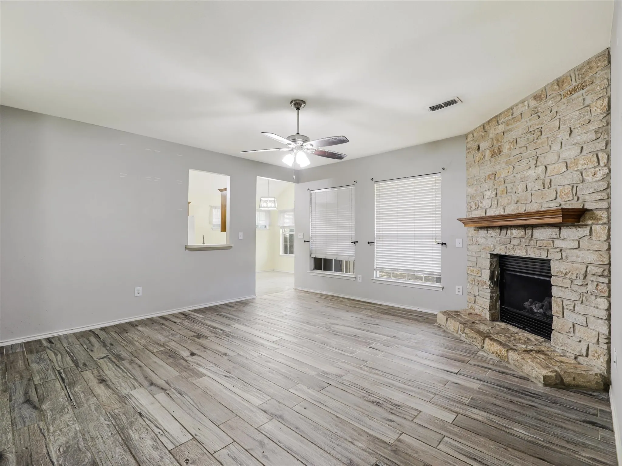 Wood-finish flooring, stone-clad fireplace with wood mantle, ceiling fan, and multiple windows with blinds