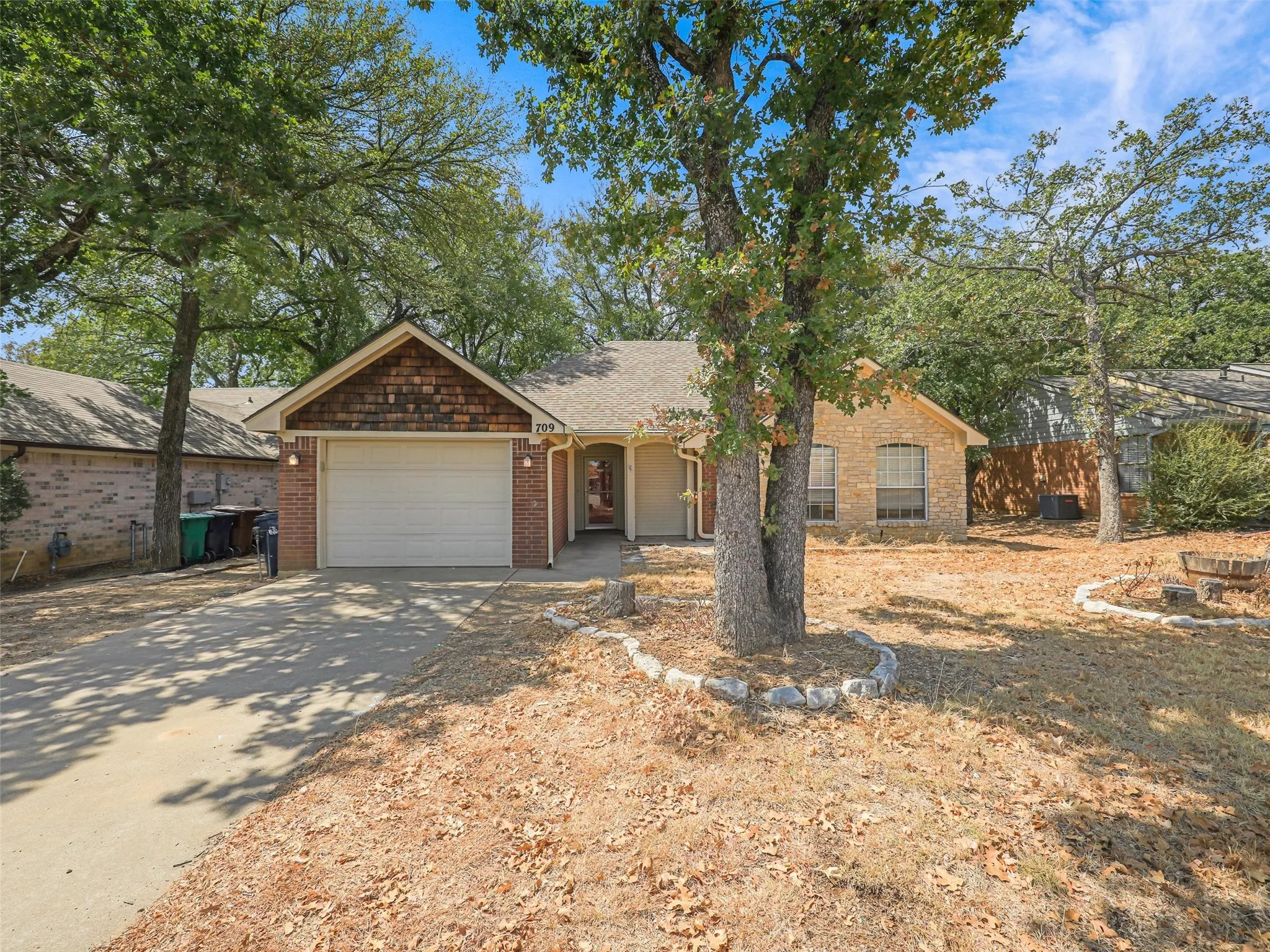 Brick exterior with natural wood shake accent, attached garage, concrete driveway, and mature trees