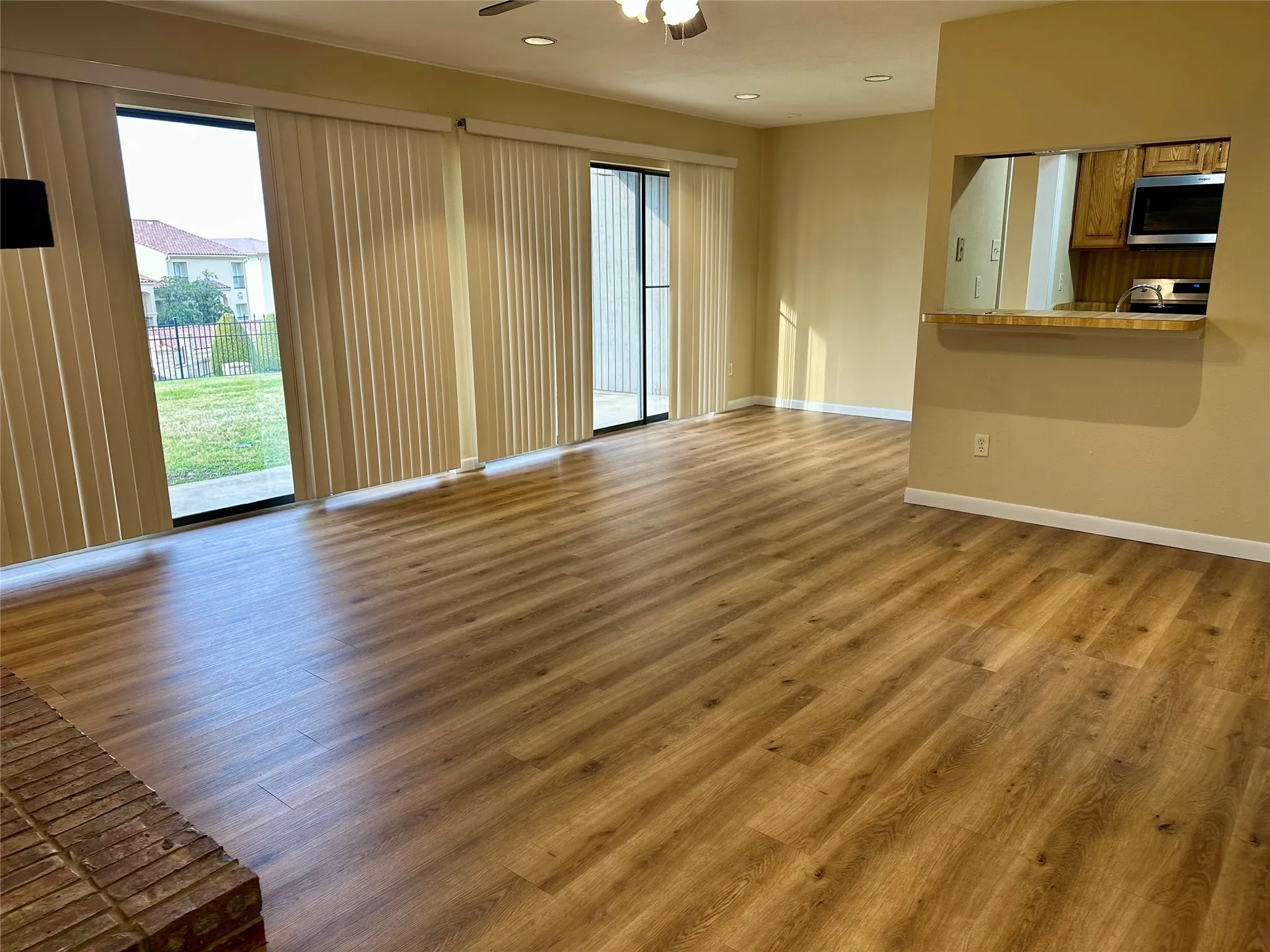 Wood-finish flooring, brick fireplace, sliding glass door with vertical blinds, recessed lighting, and a pass-through window with a breakfast bar