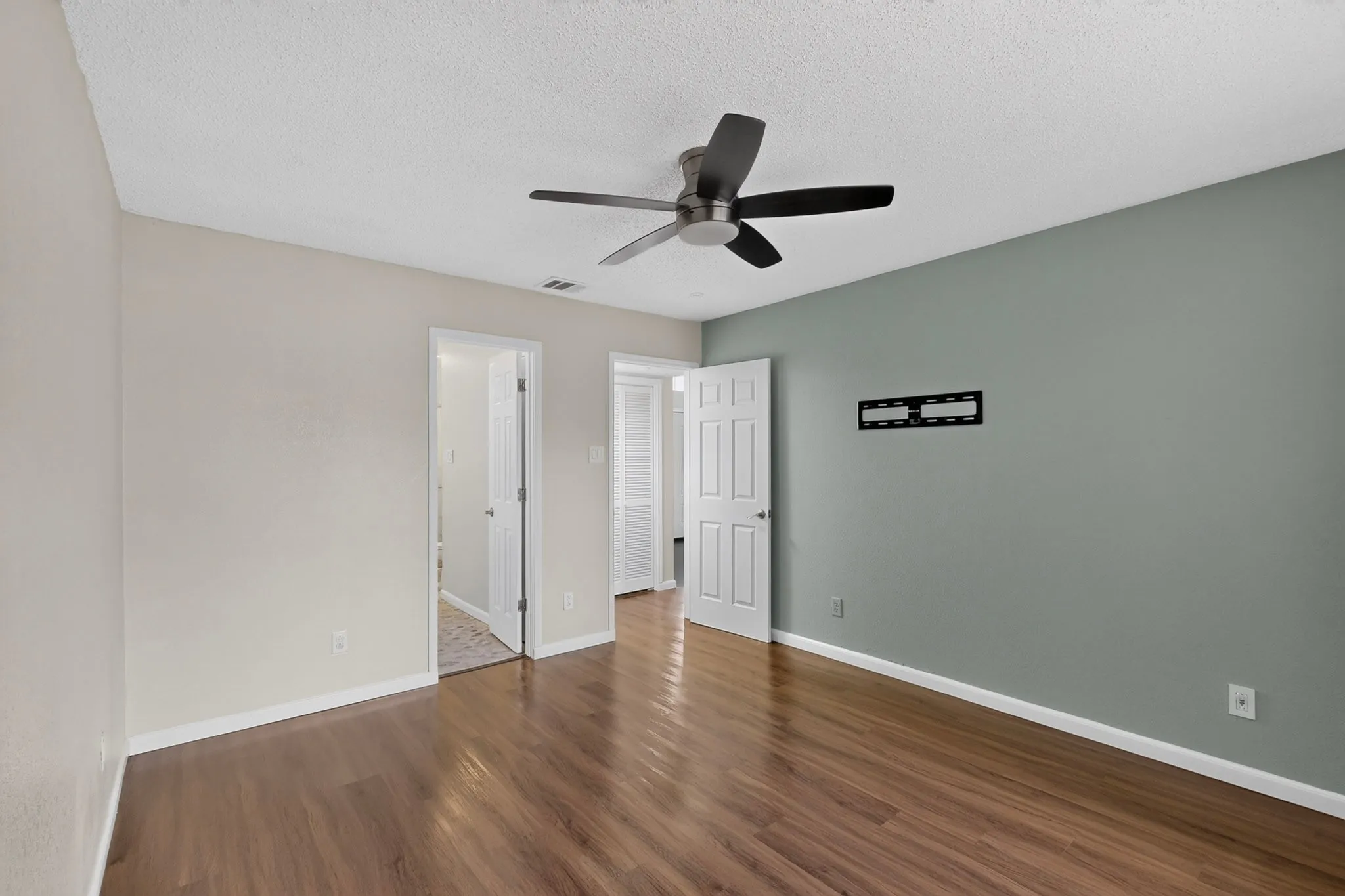 Wood-finish flooring, ceiling fan with matte black blades, white interior doors, and a wall-mounted bracket