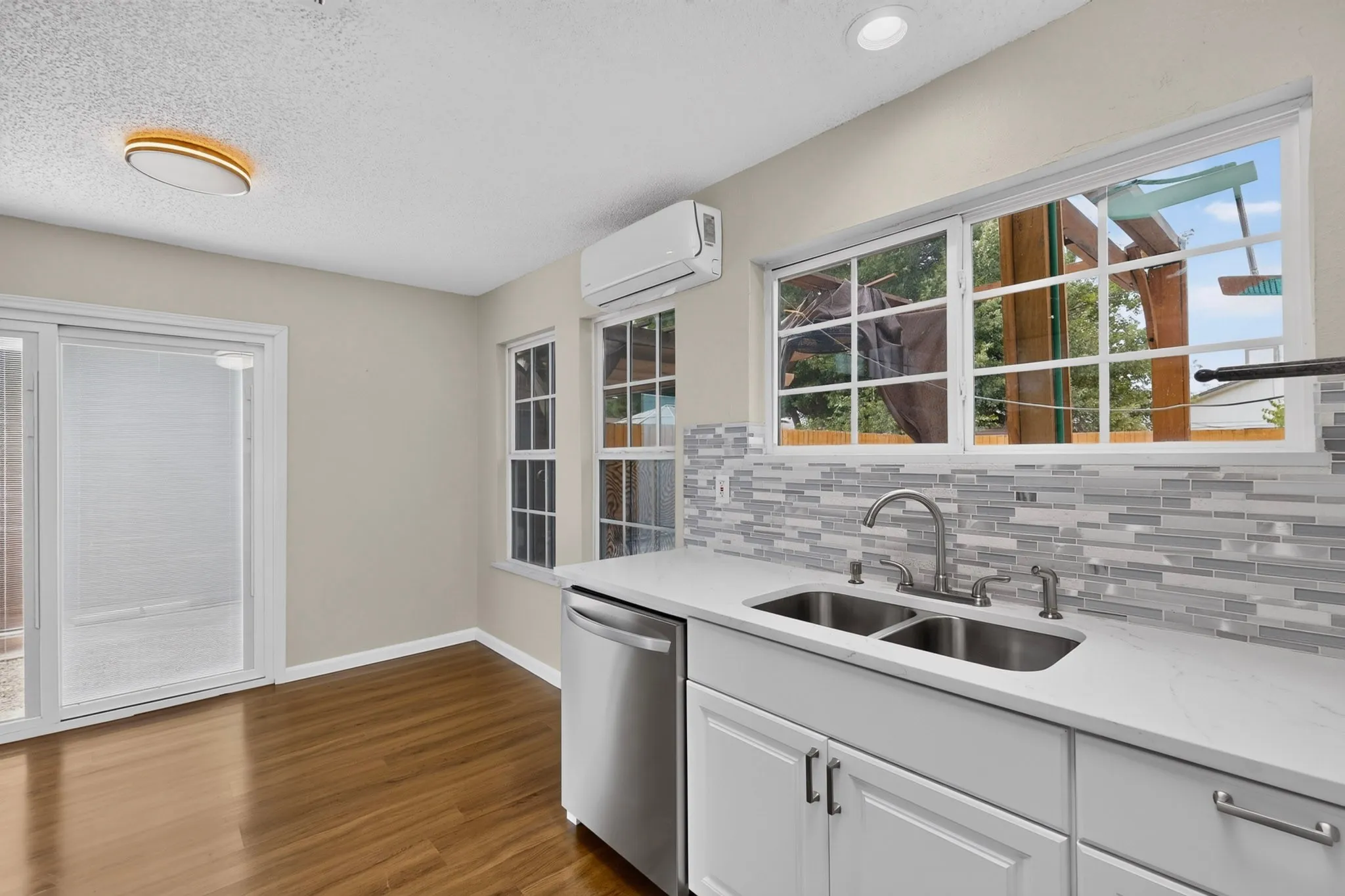 Sliding glass door, wood-finish flooring, white cabinetry, stainless steel double-basin sink, and a multi-tone tile backsplash
