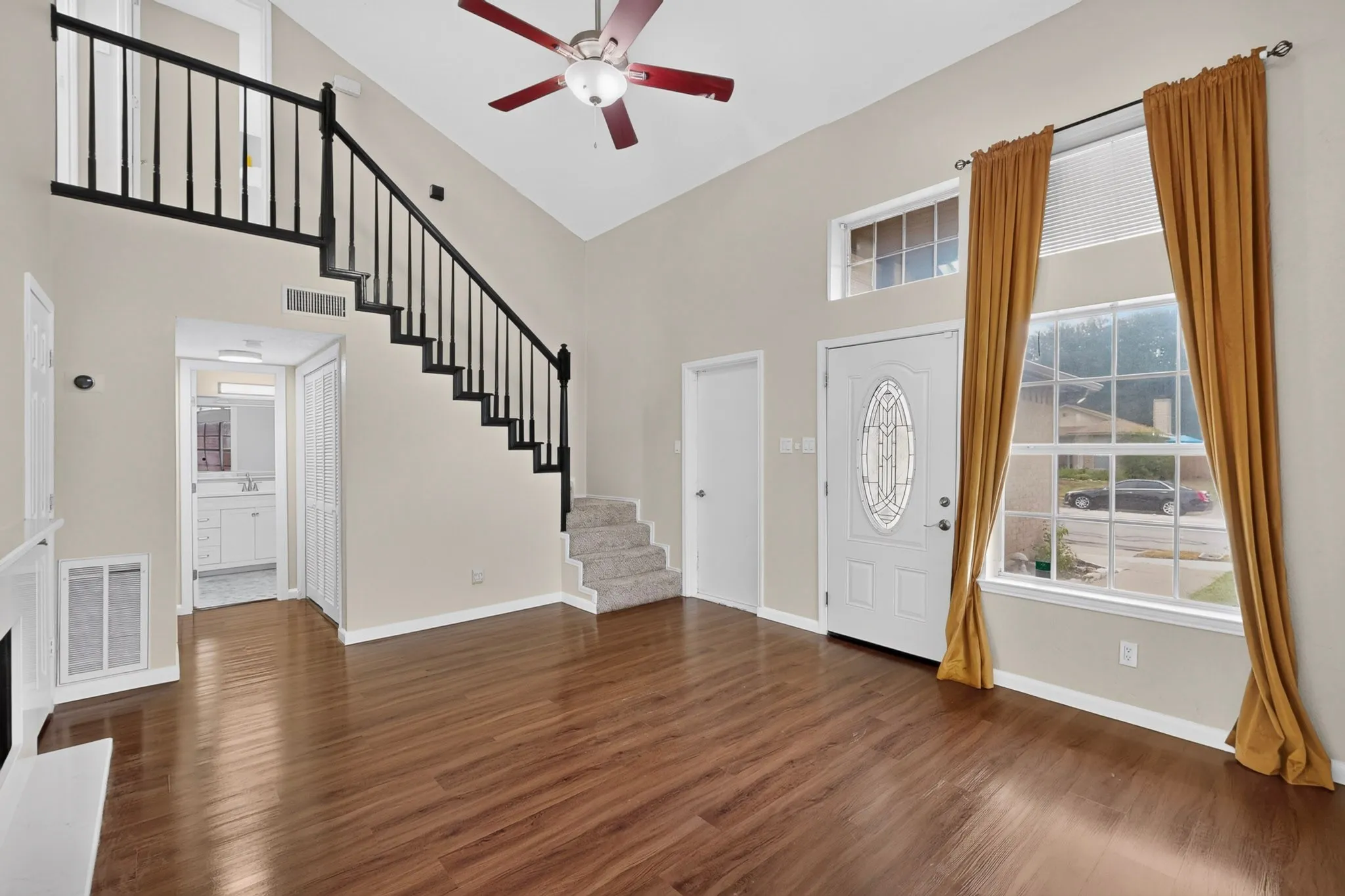 Vaulted ceiling with a ceiling fan, wood-finish flooring, staircase with black wrought iron spindles, white entry door with leaded glass insert, and a transom window