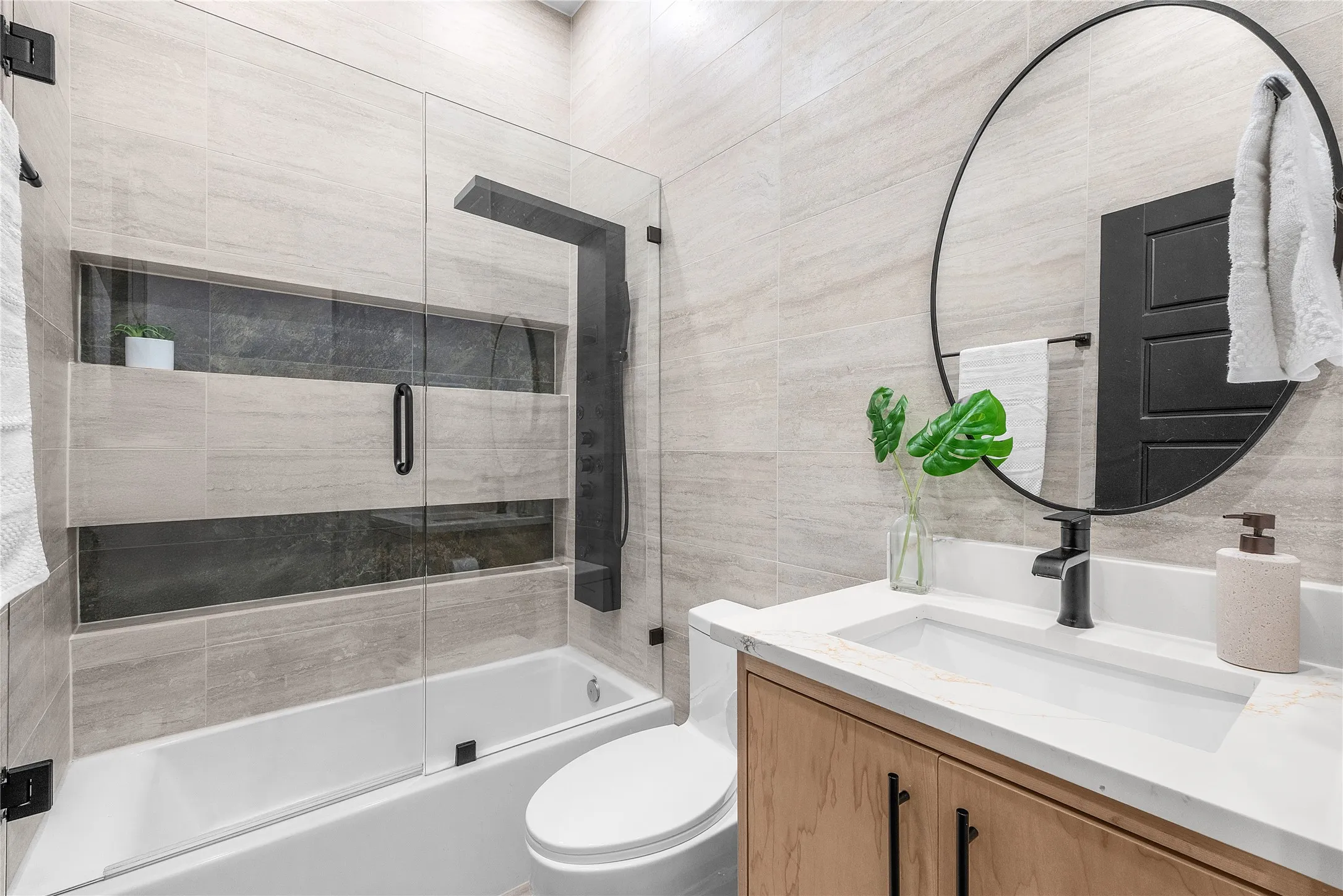 Bathtub with glass enclosure and black shower system, vanity with integrated sink, light-toned wood cabinetry, and a round black-framed mirror