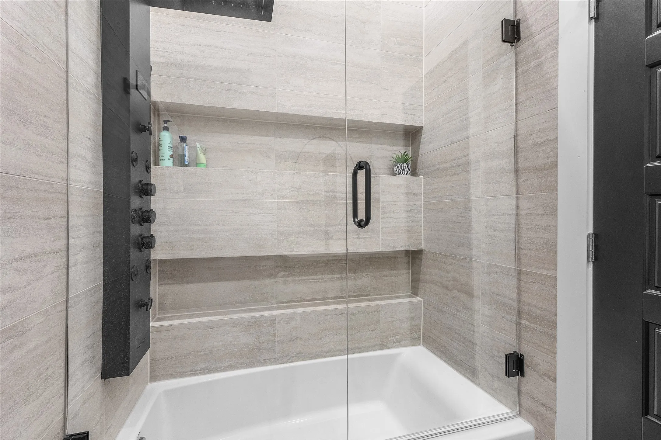 Bathtub and shower combination featuring a multi-function shower panel, light-toned tile surround, glass shower enclosure, and integrated shelving