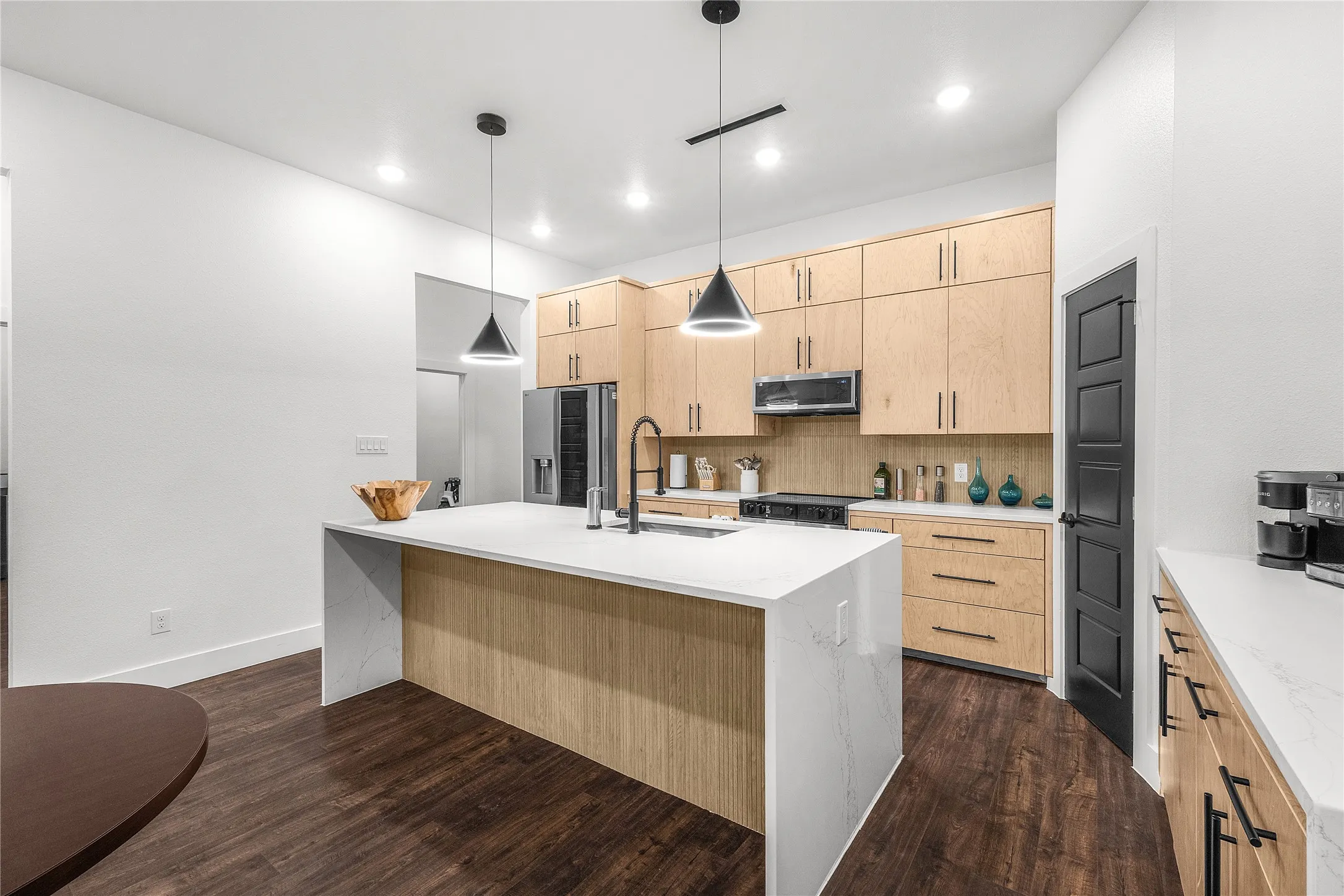 Modern kitchen featuring light wood-finish cabinetry, dark wood-finish flooring, a white countertop island, stainless steel appliances, and contemporary pendant lighting
