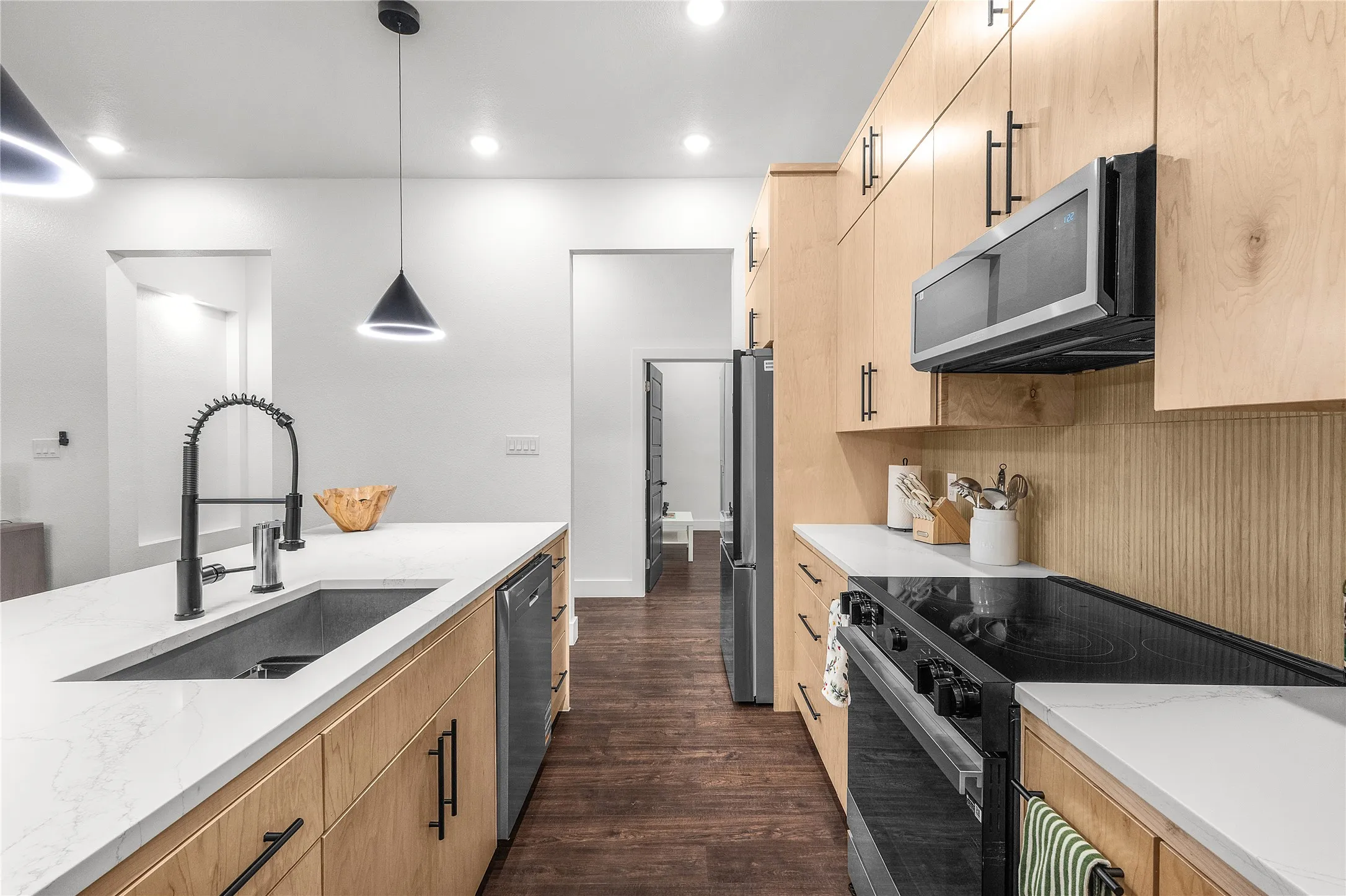 Contemporary kitchen featuring light wood-finish cabinetry, stainless steel appliances, a center island with a large sink, and dark wood-finish flooring