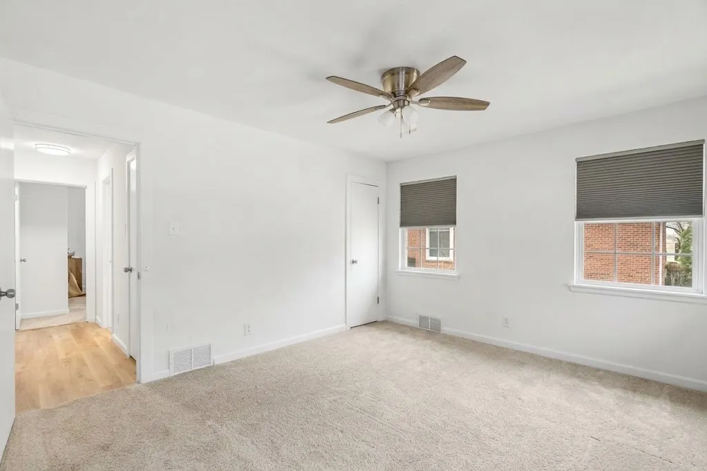 Neutral wall tones, recessed ceiling lighting, light-colored carpeting, two windows with blinds, and a ceiling fan with integrated lighting