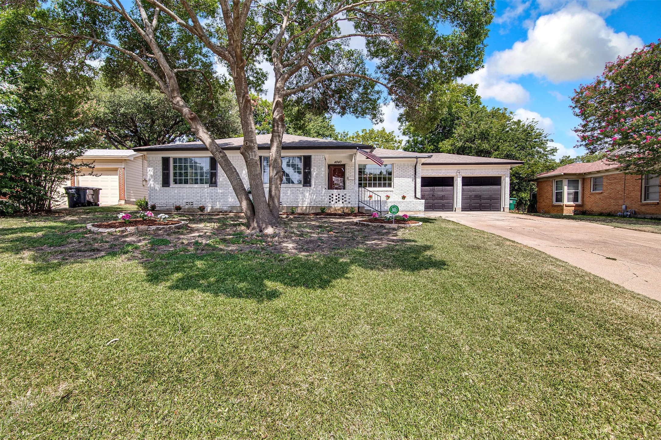 White brick exterior with black shutters, mature tree, two-car garage, and concrete driveway
