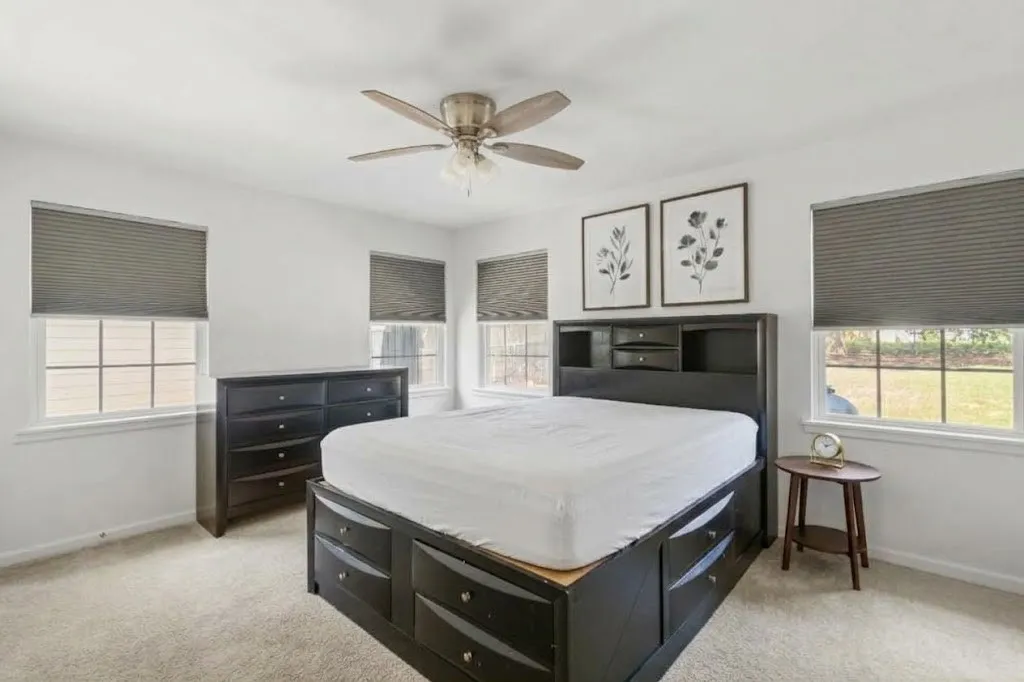 Light-colored carpeting, multiple windows with blinds, and a ceiling fan with integrated lighting