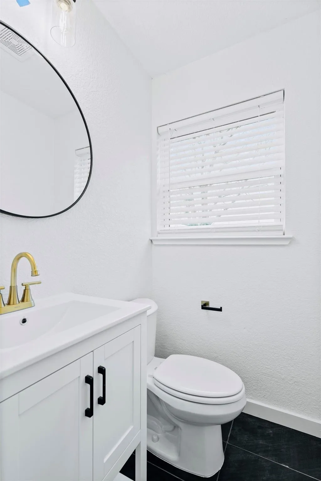 White vanity with black hardware, gold-finish faucet, dark tile flooring, and a contemporary oval mirror