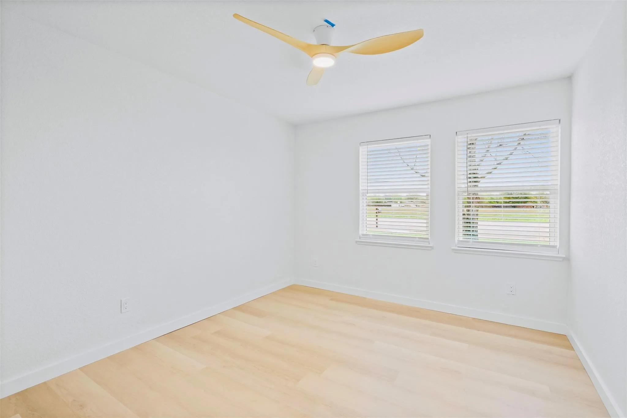 Light wood-finish flooring, white walls, two windows with blinds, and a modern ceiling fan with integrated lighting
