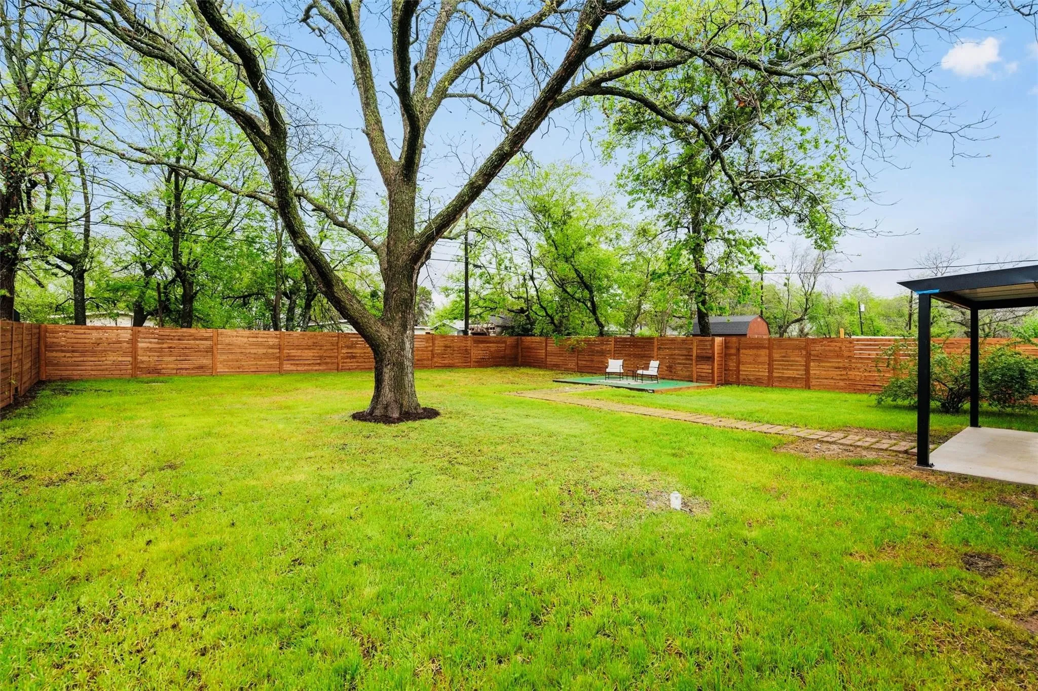 Spacious backyard featuring a large tree, manicured lawn, wood fencing, and a covered patio area with concrete flooring