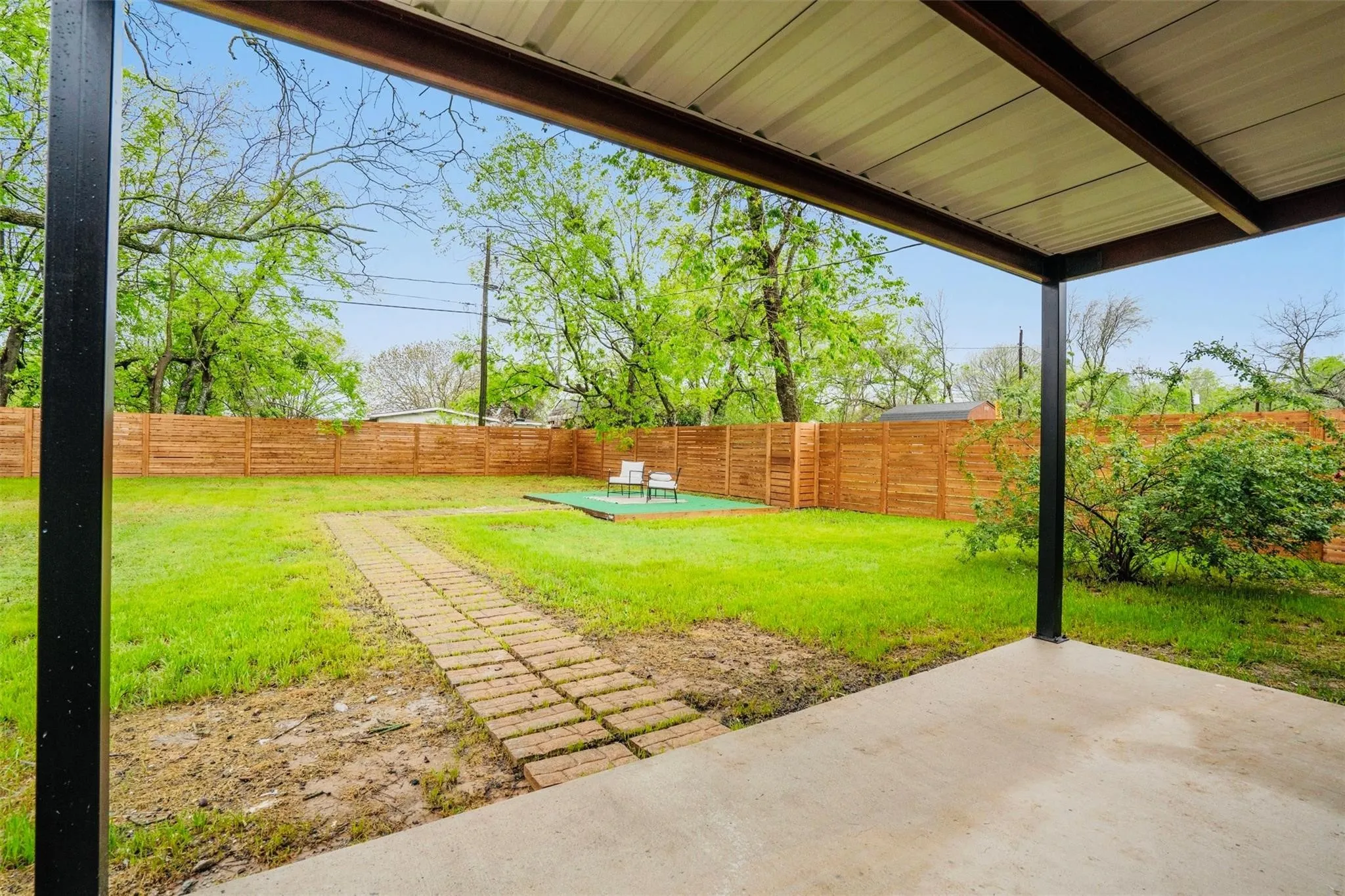 Covered patio with concrete flooring, extending to a fenced yard with a brick paver pathway and natural landscaping