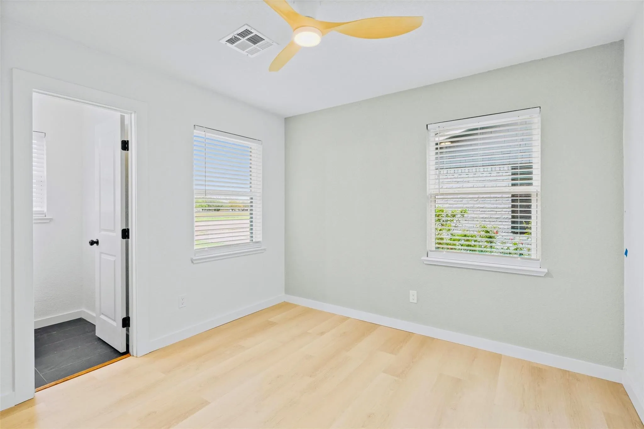 Light wood-finish flooring, two windows with blinds, a ceiling fan with integrated lighting, and a white door with dark hardware