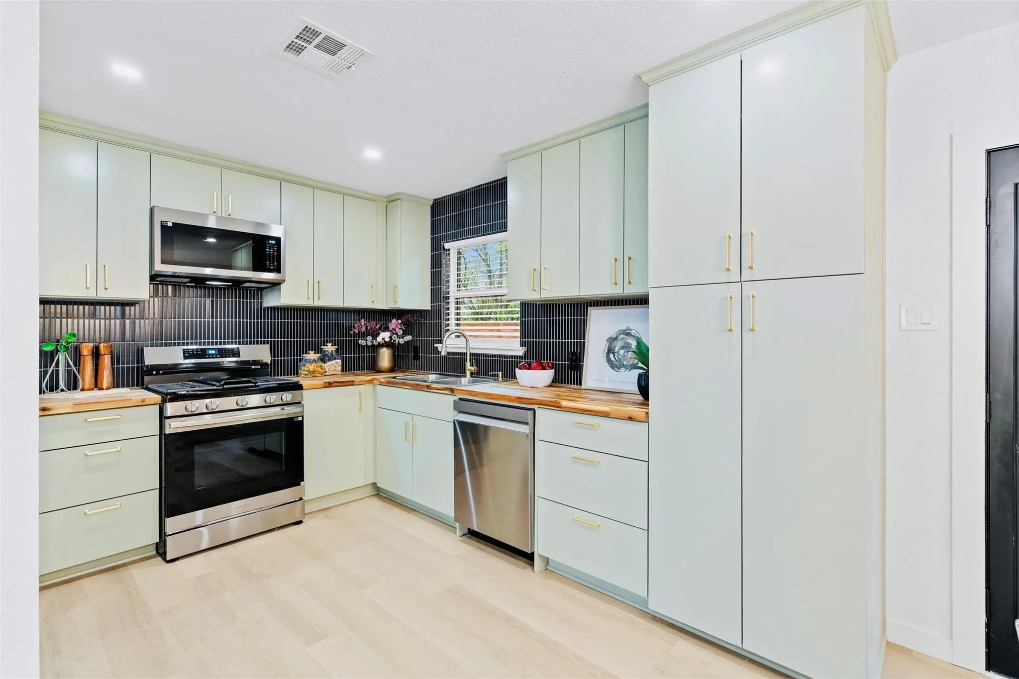 Modern kitchen featuring light-toned cabinetry with gold-finish hardware, wood-finish countertops, stainless steel appliances, and a black tile backsplash