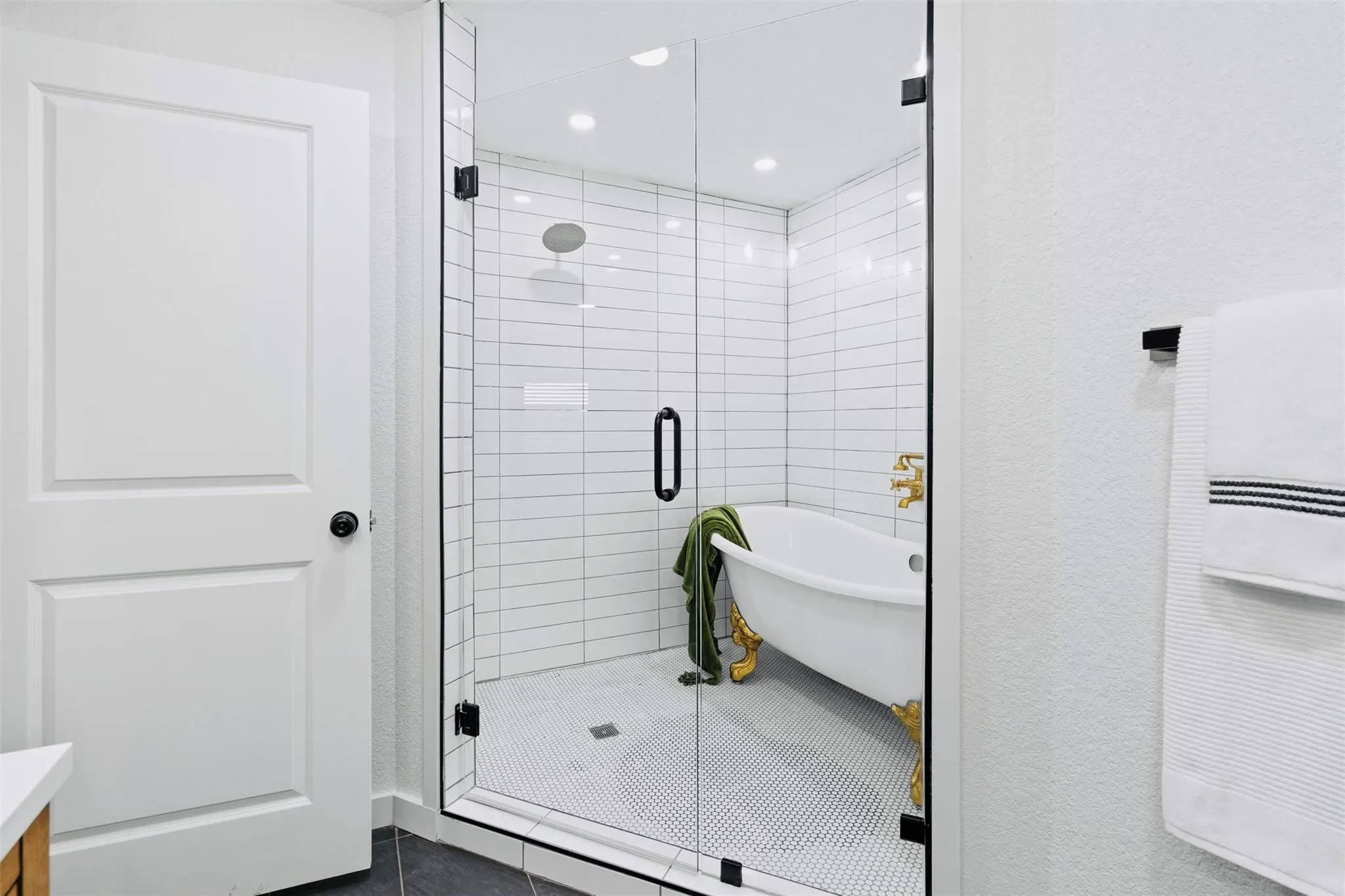 Frameless glass shower enclosure with matte black hardware, white subway tile walls, and a freestanding clawfoot tub with gold-tone feet and fixtures