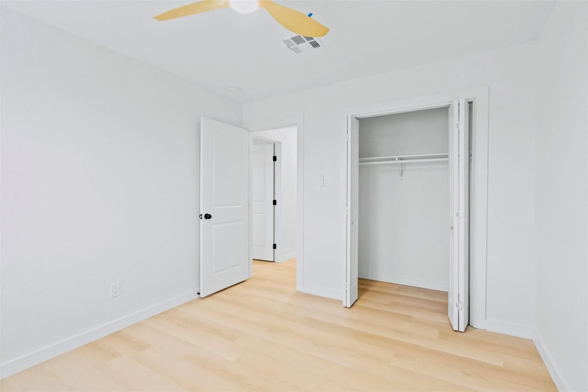 Light wood-finish flooring, white walls, ceiling fan, bi-fold closet door, and interior paneled door with dark hardware