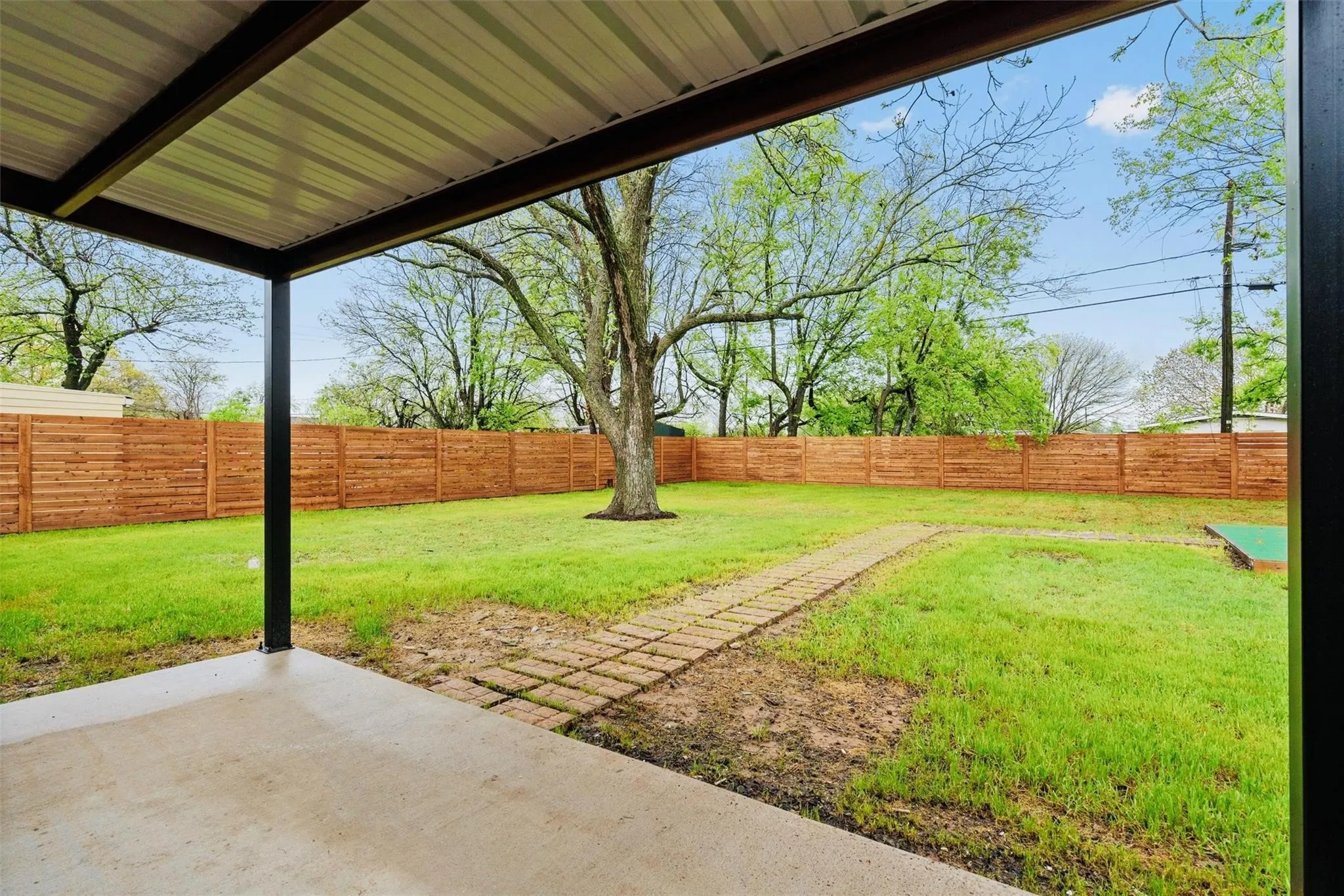 Covered patio with concrete flooring, framed by a wood horizontal plank fence, and a grass lawn with a paver pathway