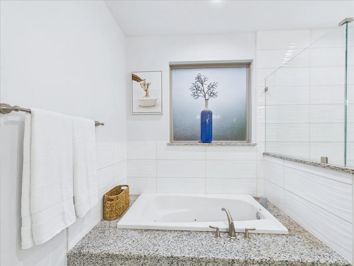 Built-in bathtub with deck-mounted faucet, surrounded by a speckled countertop and white tiled walls