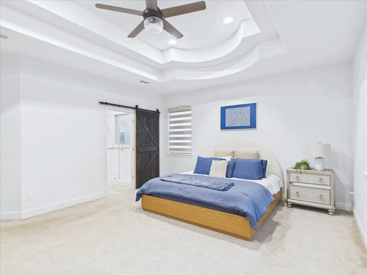 Tray ceiling with recessed lighting and a ceiling fan, light-toned carpeting, and a dark wood-finish barn door