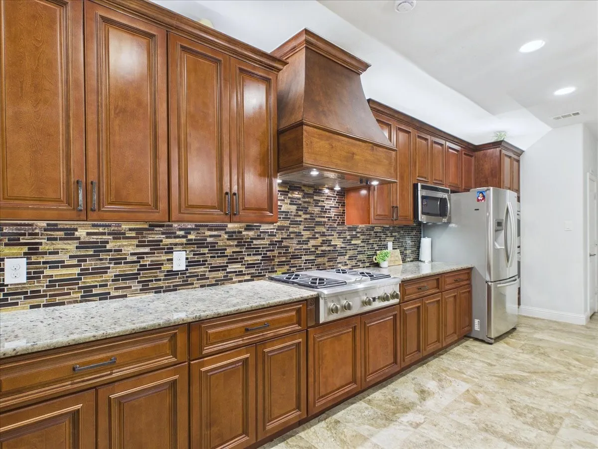 Kitchen featuring wood cabinetry, an integrated wood range hood, a mosaic tile backsplash, light-toned countertops, and stainless steel appliances