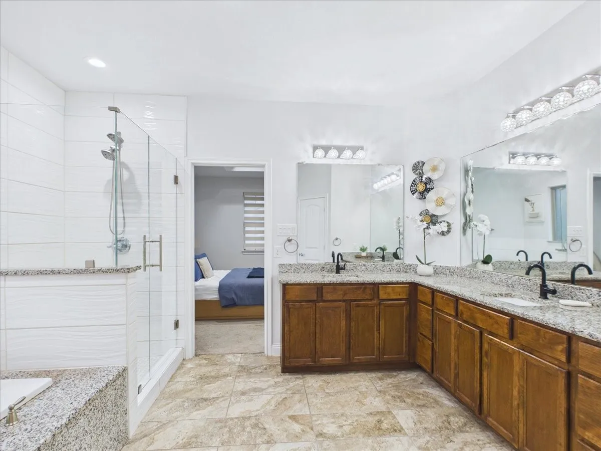 Spacious bathroom featuring a glass-enclosed shower, dual vanities with granite countertops, wood-finish cabinetry, and large format floor tiling