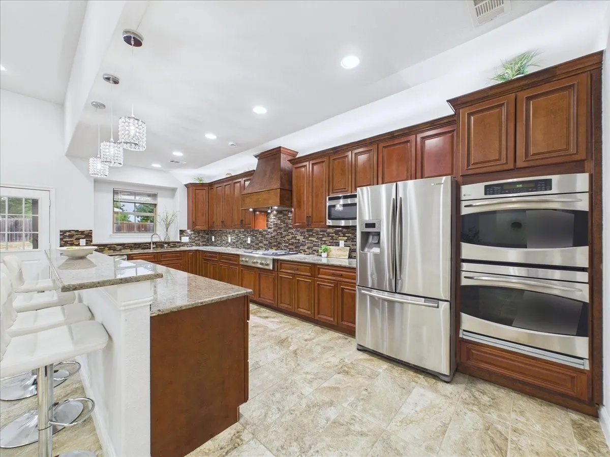 Kitchen featuring wood cabinetry, stainless steel appliances, a multi-tone tile backsplash, granite countertops, and light-toned tile flooring