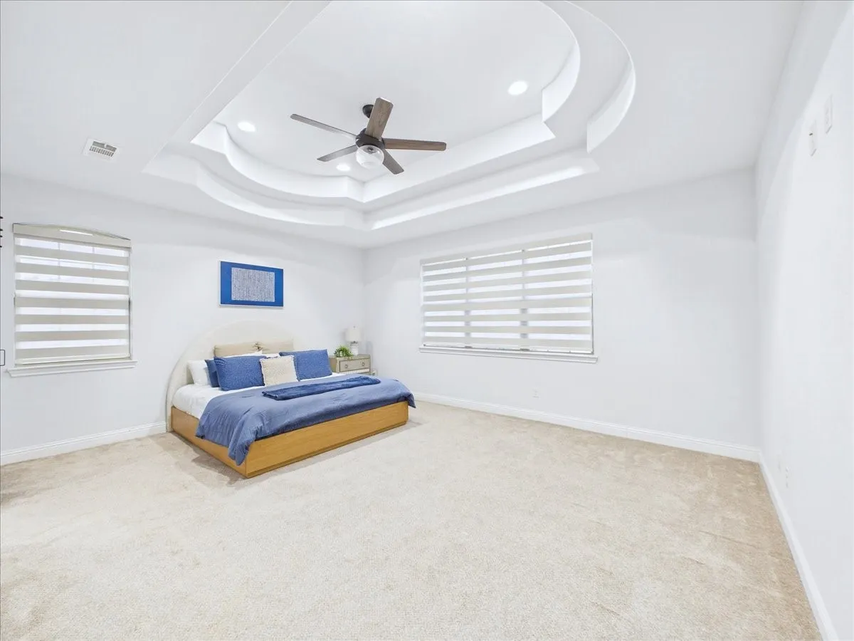 Tray ceiling with recessed lighting and a ceiling fan, dual windows with blinds, and light-colored carpeting