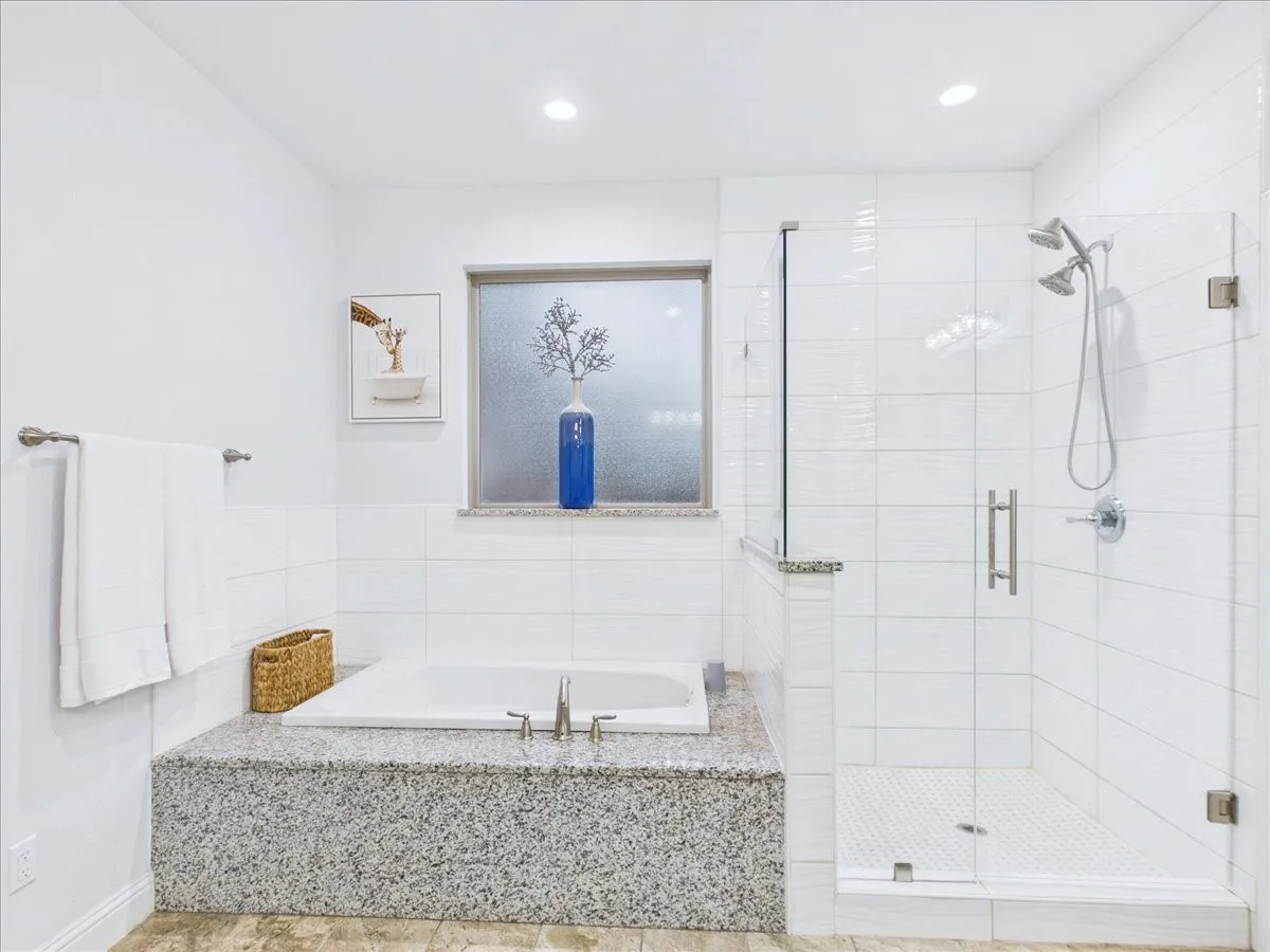 Soaking tub with a speckled stone surround, glass-enclosed shower, dual shower heads, and white tiled walls