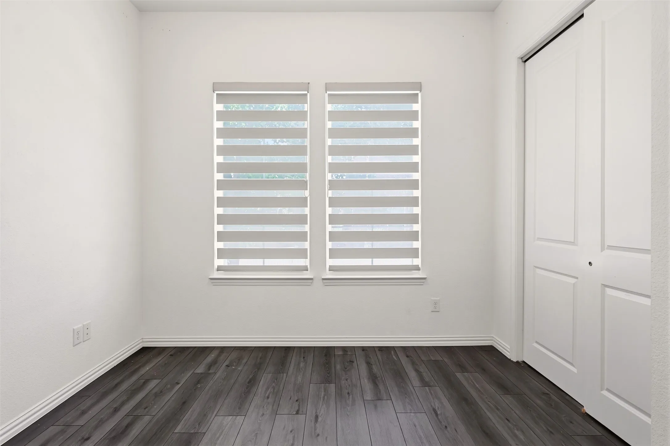Room featuring dual windows with blinds, wood-finish flooring, white baseboards, and a bi-fold closet door