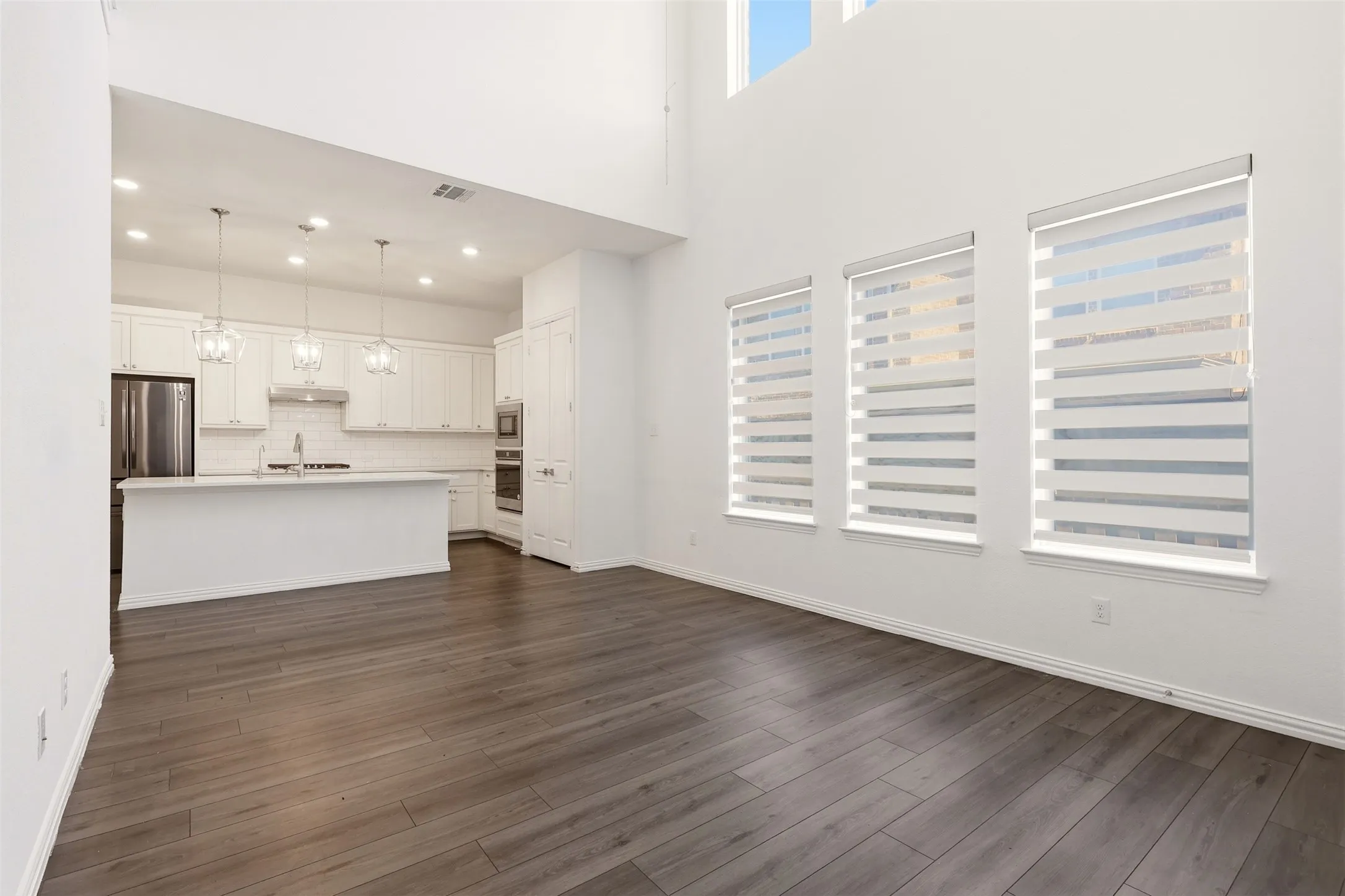 Open-concept living area featuring high ceilings, wood-finish flooring, and multiple windows with layered privacy blinds