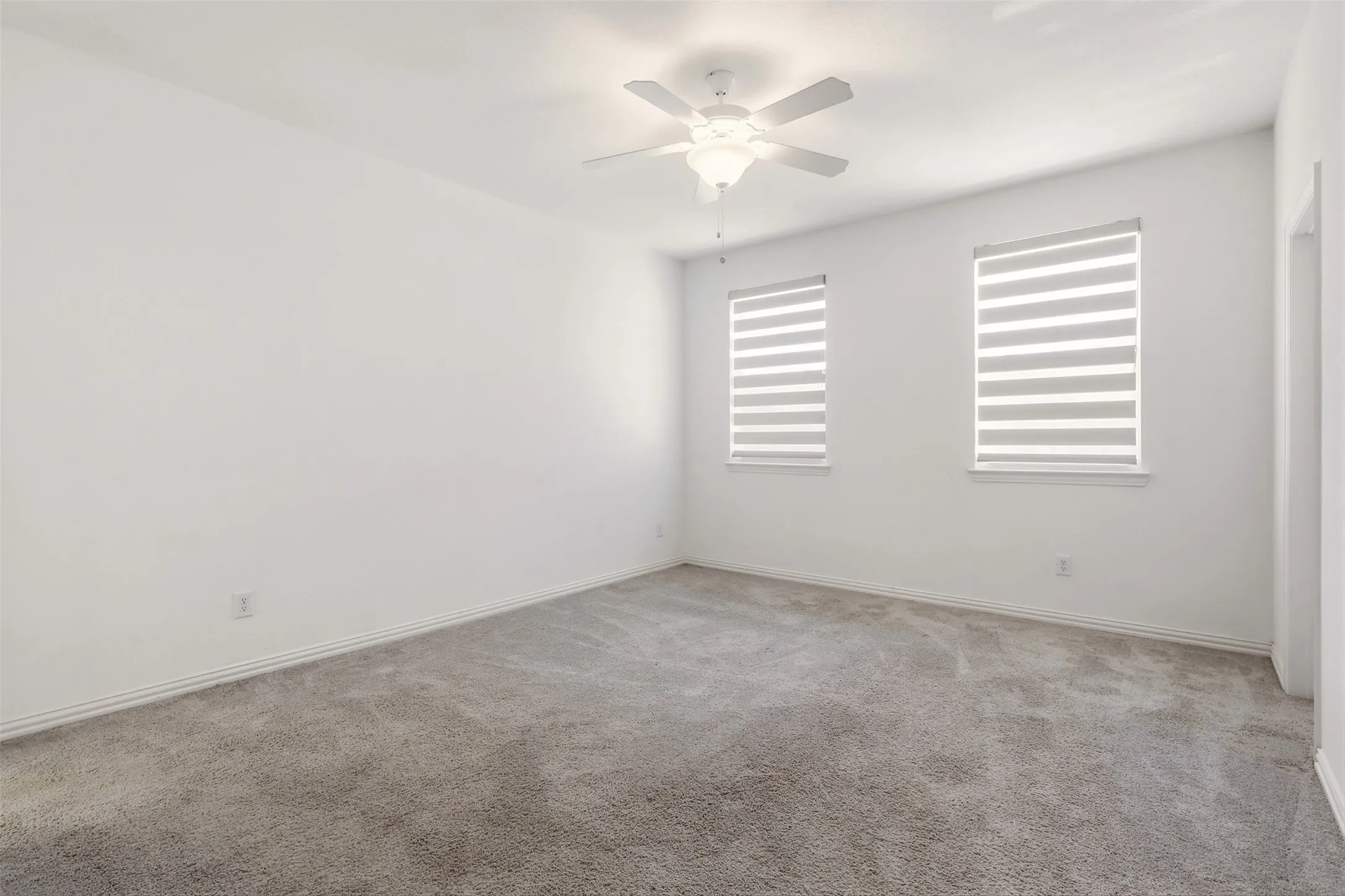 Spacious room featuring neutral carpeting, white walls, two windows with horizontal blinds, a ceiling fan with integrated lighting, and white baseboards