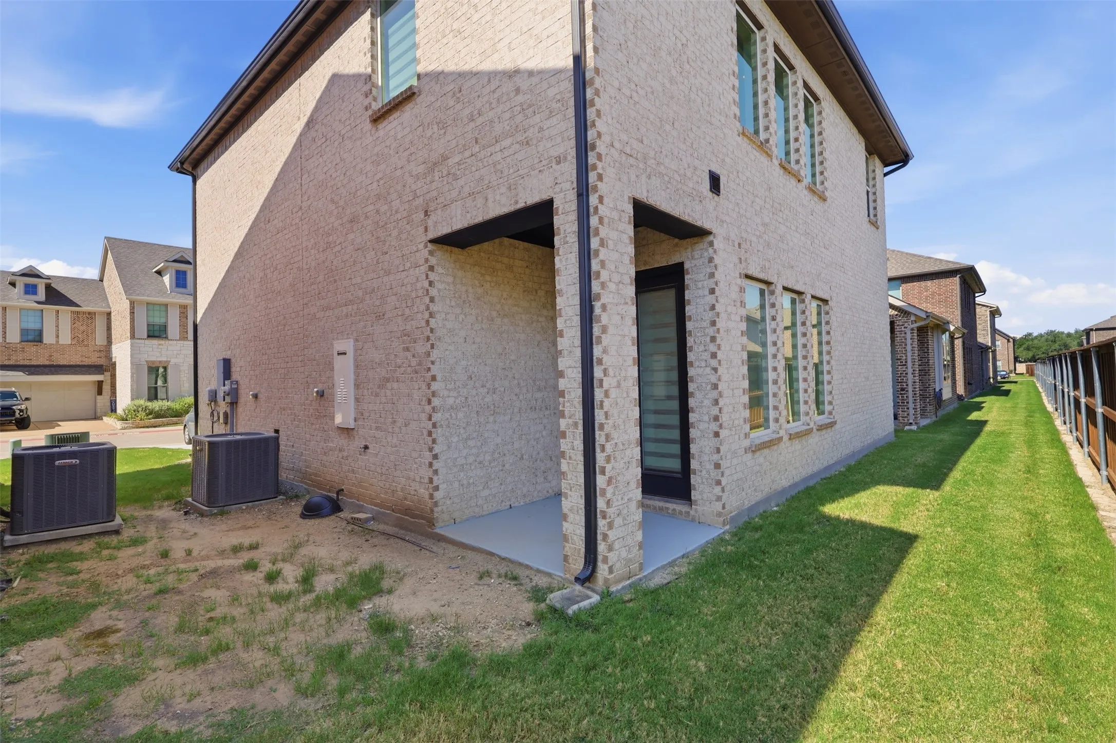 Two-story exterior featuring light-colored brick, multiple windows with white frames, a covered patio area with a concrete slab, and a narrow grassy side yard