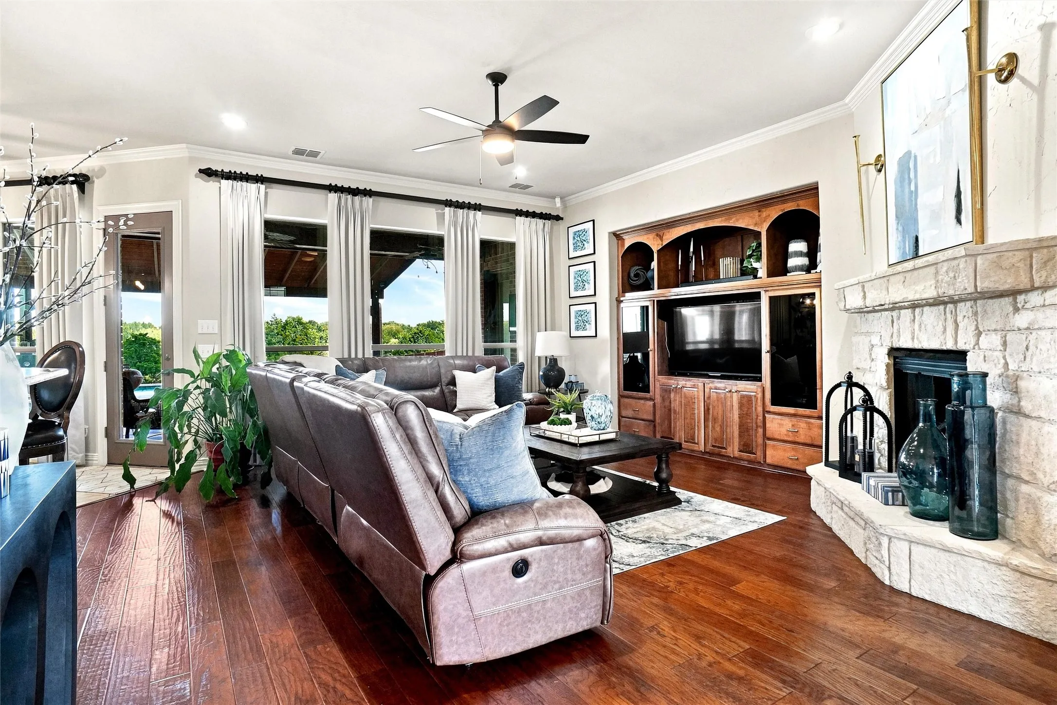 Wood-finish flooring throughout, featuring a stone-clad corner fireplace, a built-in media center with integrated shelving, and multiple large windows