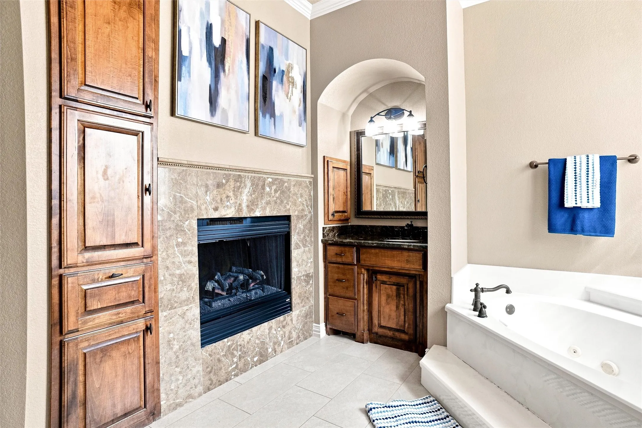 Built-in fireplace with a stone surround, white soaking tub, light-toned floor tiling, and wood cabinetry