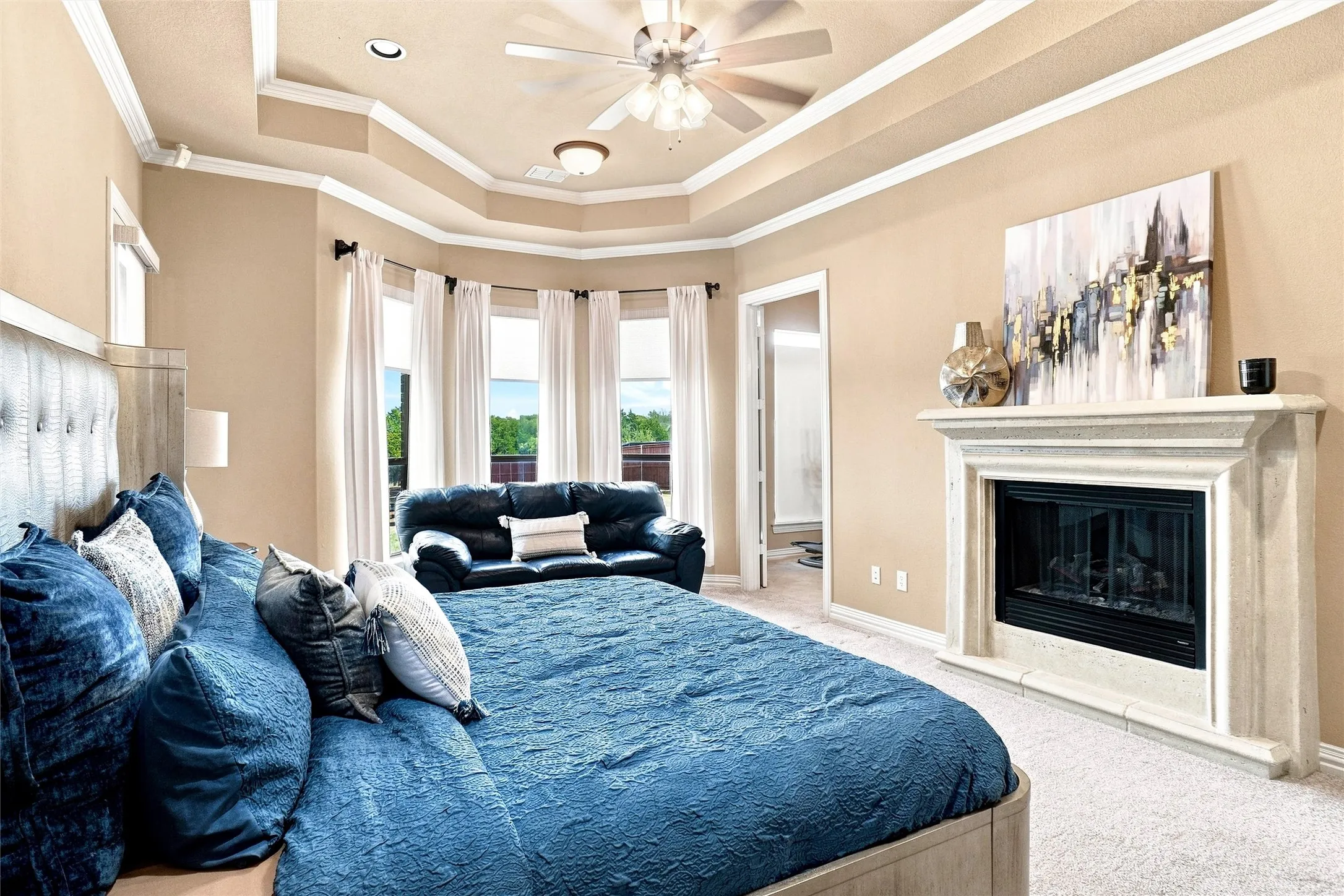 Tray ceiling with crown molding, ceiling fan, bay window, and a built-in fireplace with a stone surround